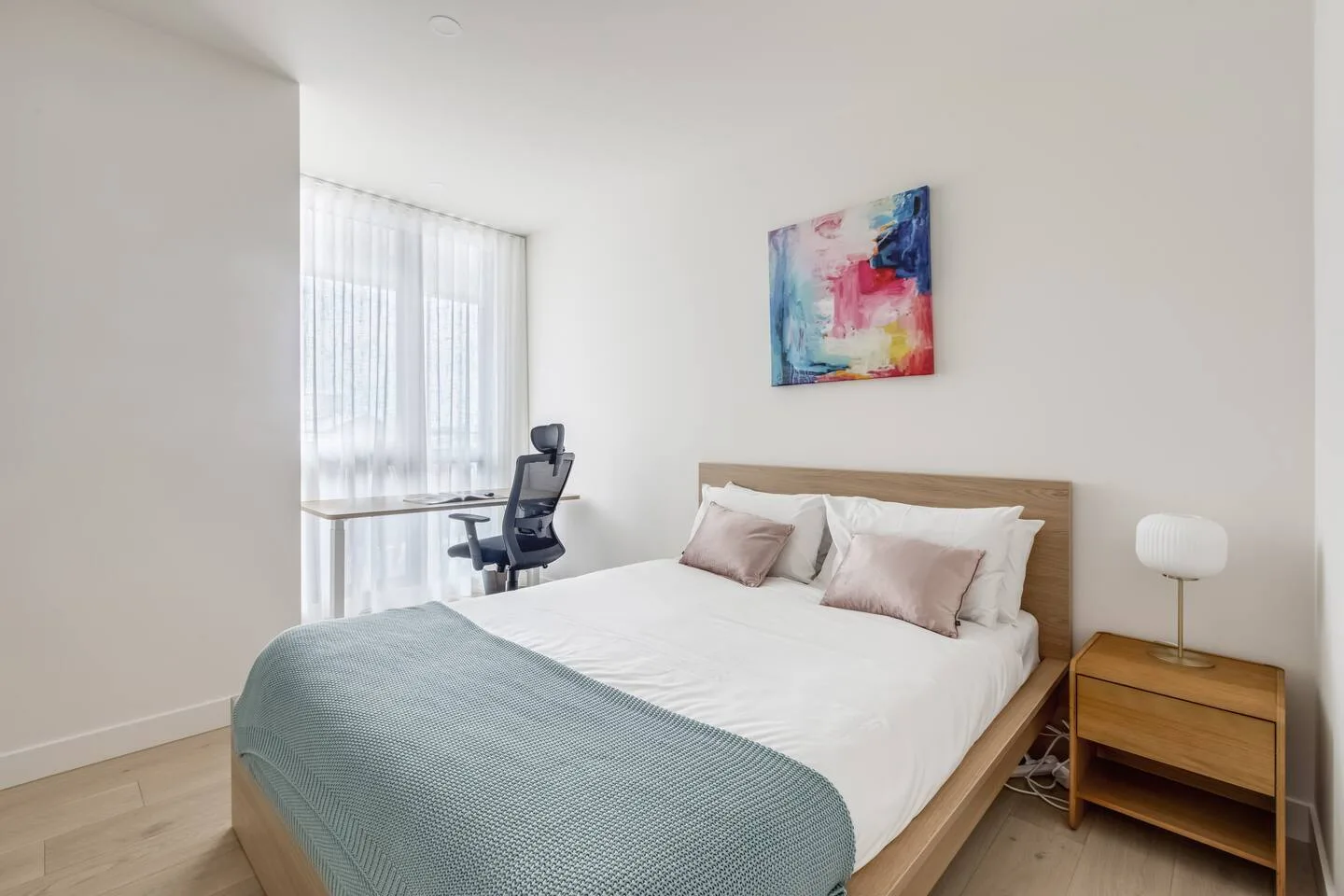 Un dormitorio luminoso tiene una cama, un escritorio junto a la ventana y arte colorido en la pared.
