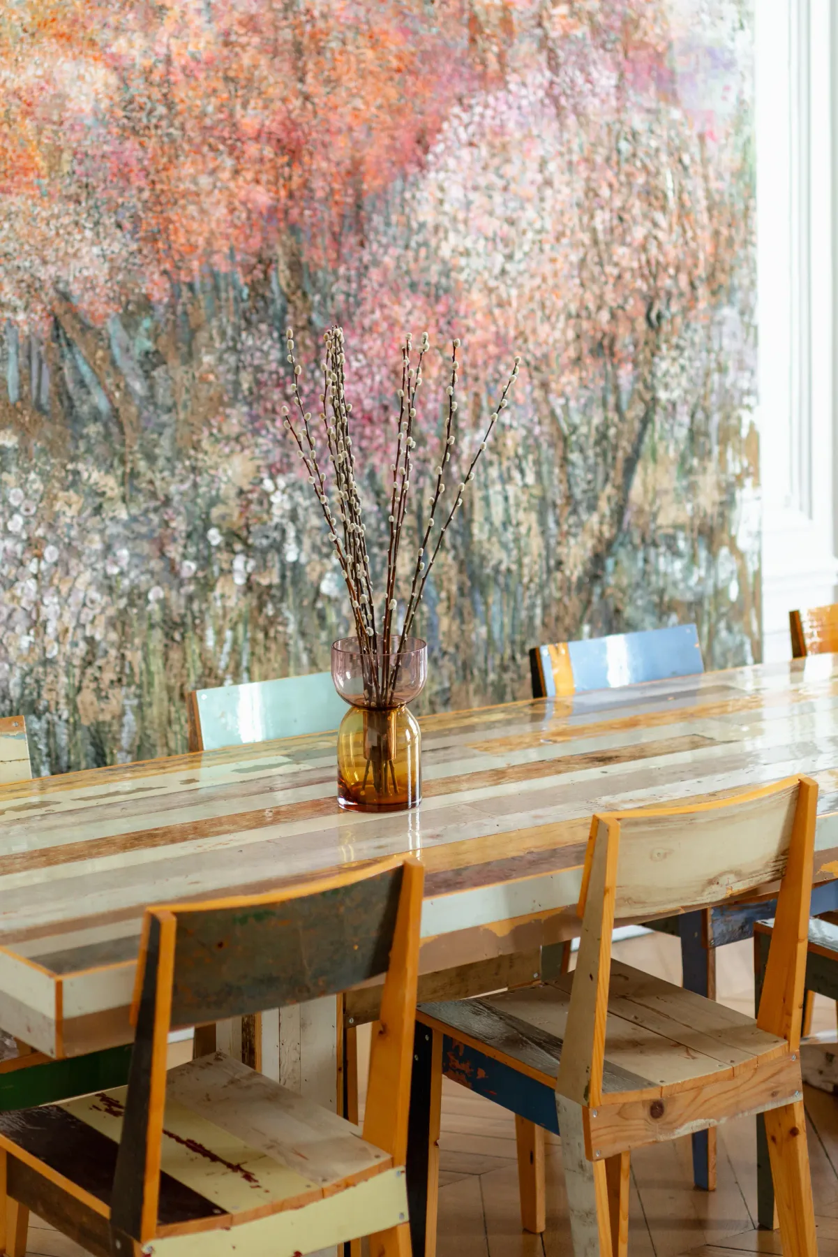 A dining area with a long table and chairs beneath a large abstract wall painting.