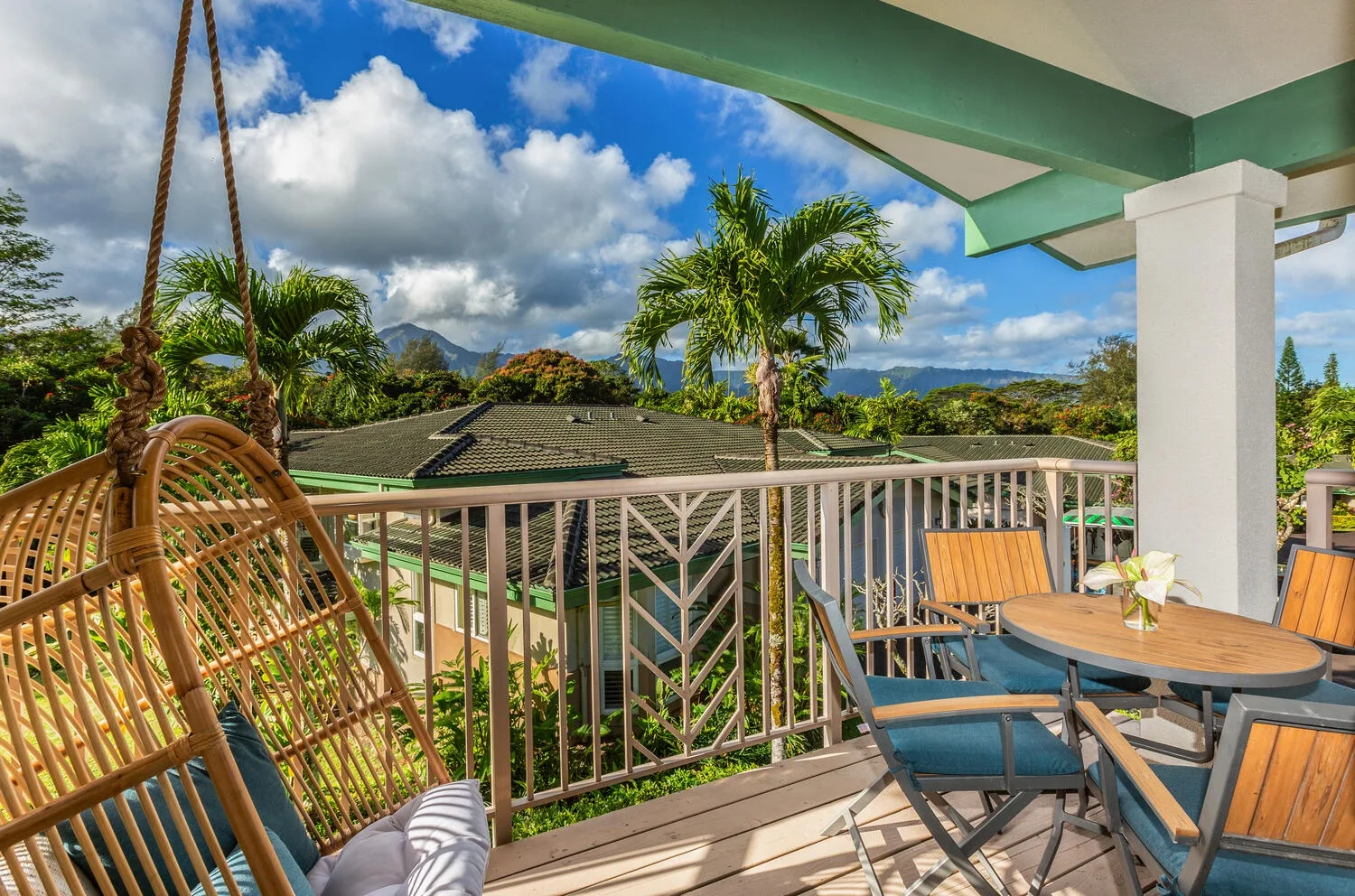 A covered outdoor balcony has a dining table and seating overlooking palm trees and greenery.