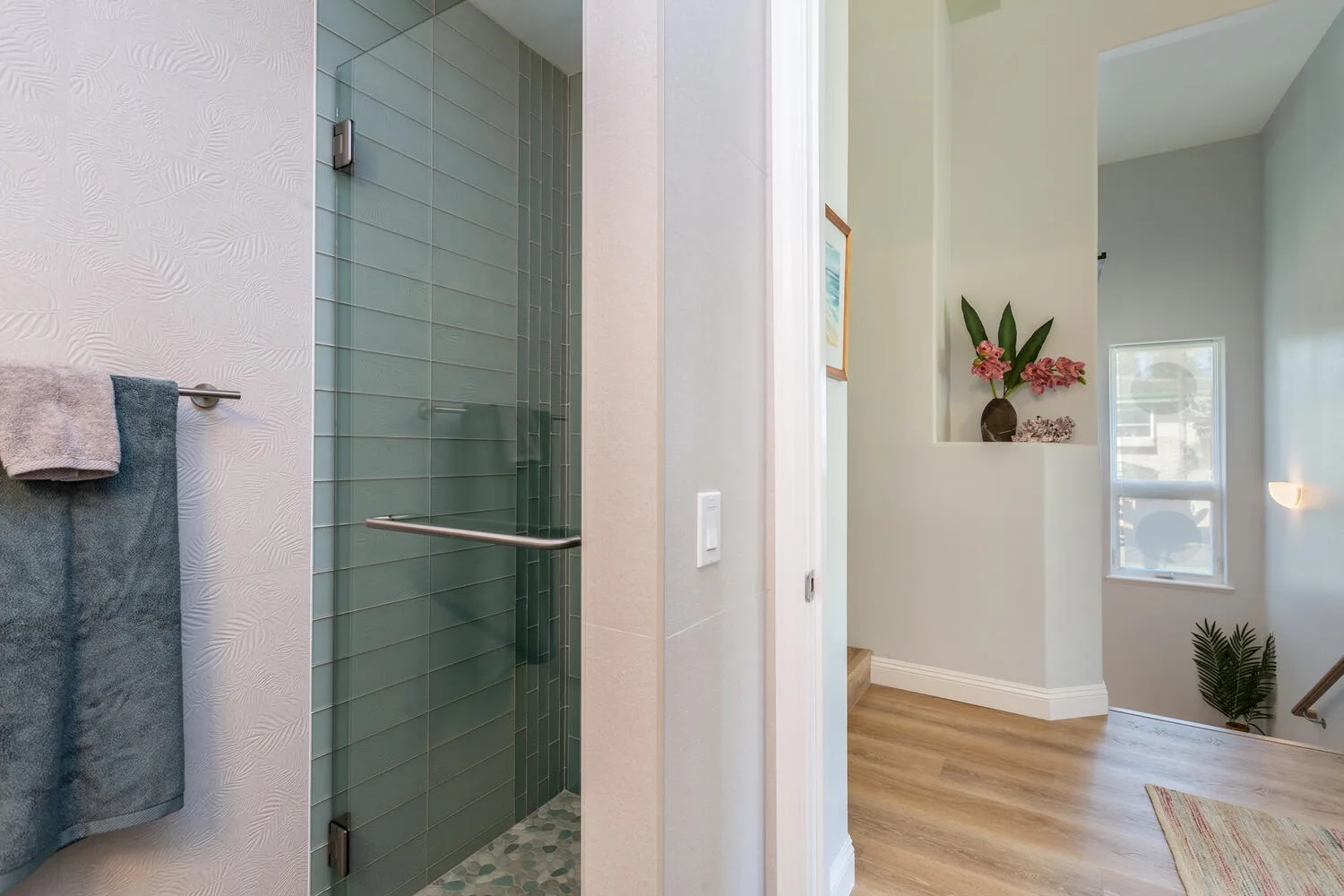 A bathroom opens onto a bright hallway with wood flooring and a window.