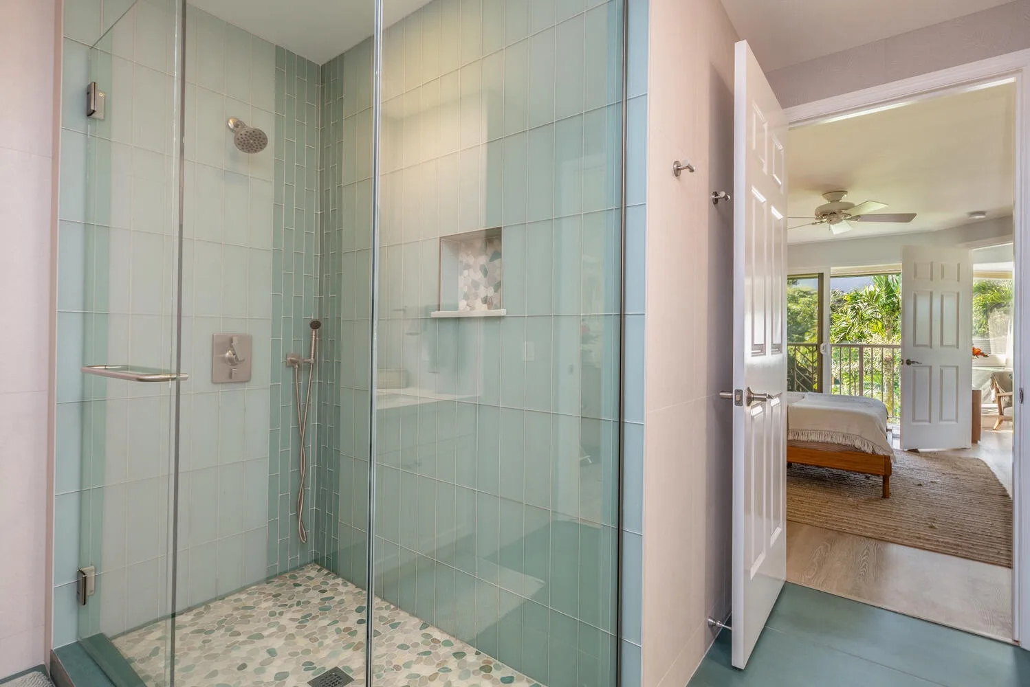 A glass-enclosed shower opens onto a bedroom with a bed and ceiling fan.