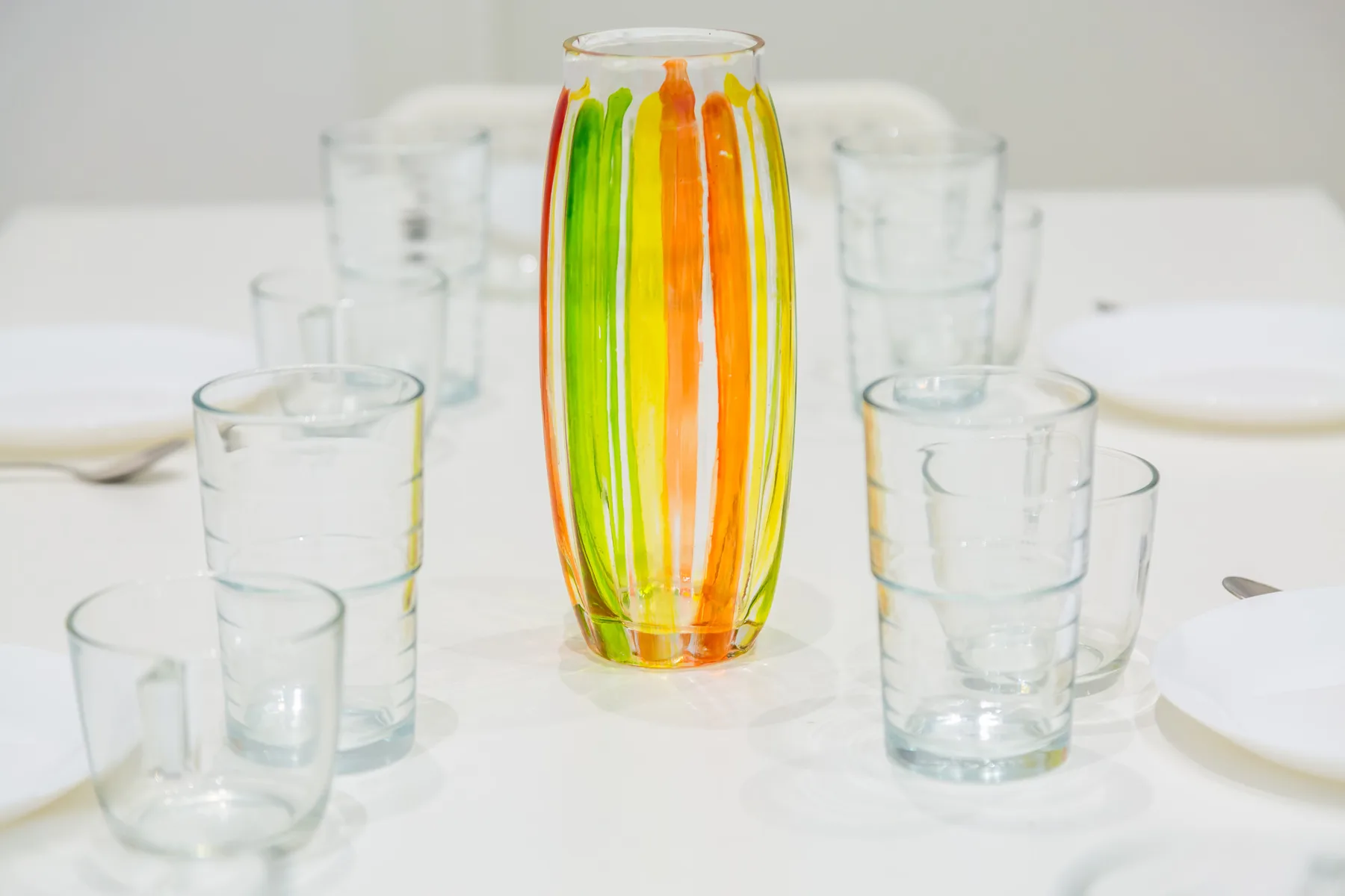 A colorful glass vase stands among clear drinking glasses on a white table.