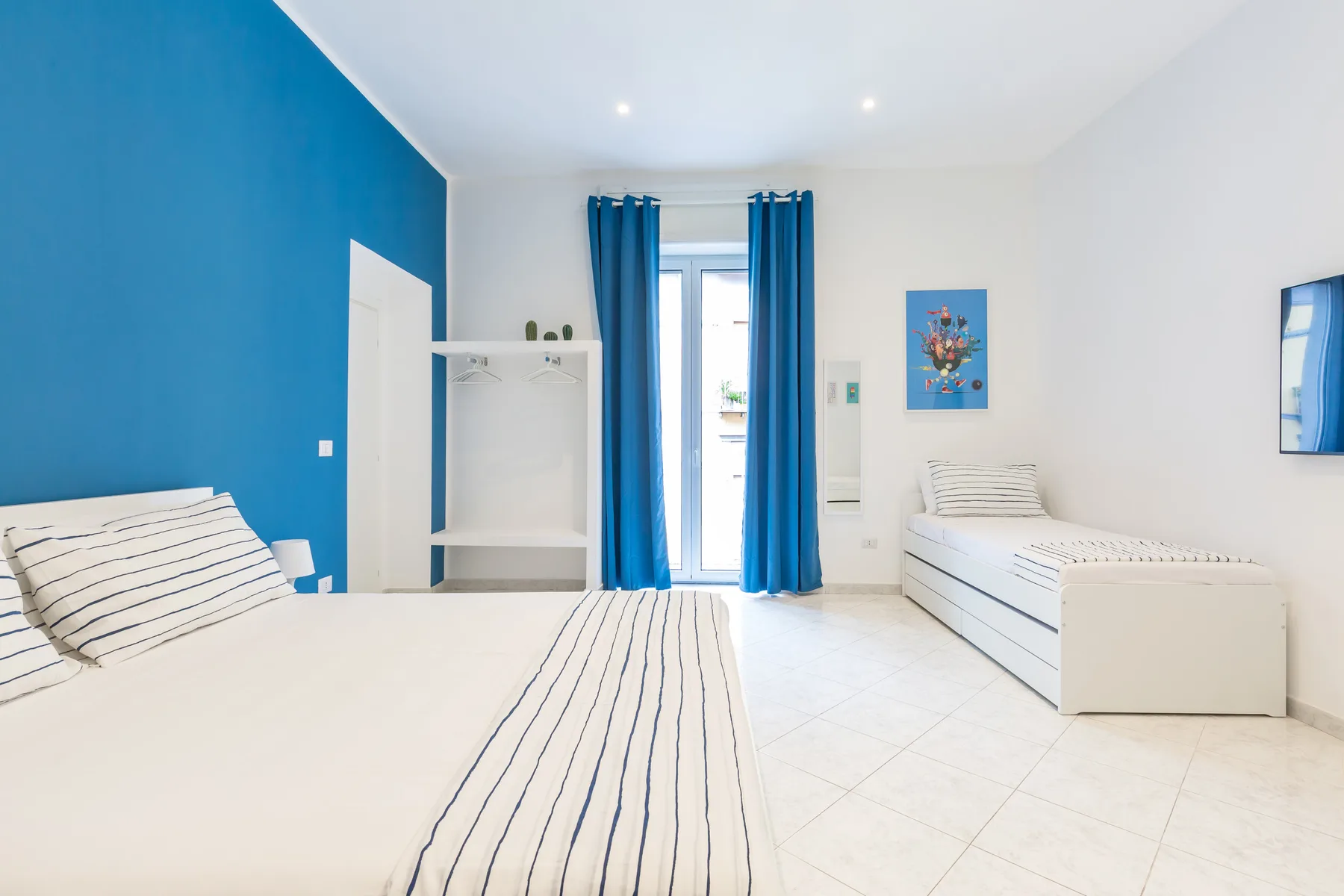 A bright bedroom with two beds and blue curtains beside a glass door.