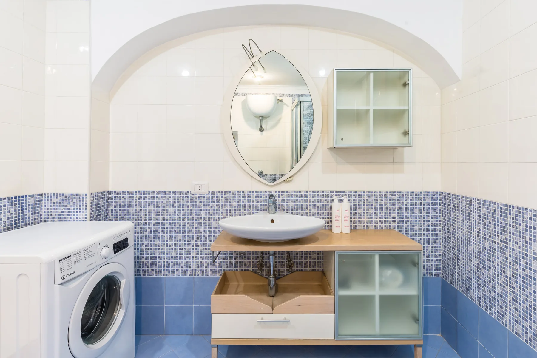 A tiled bathroom with a washing machine, sink, and round mirror.