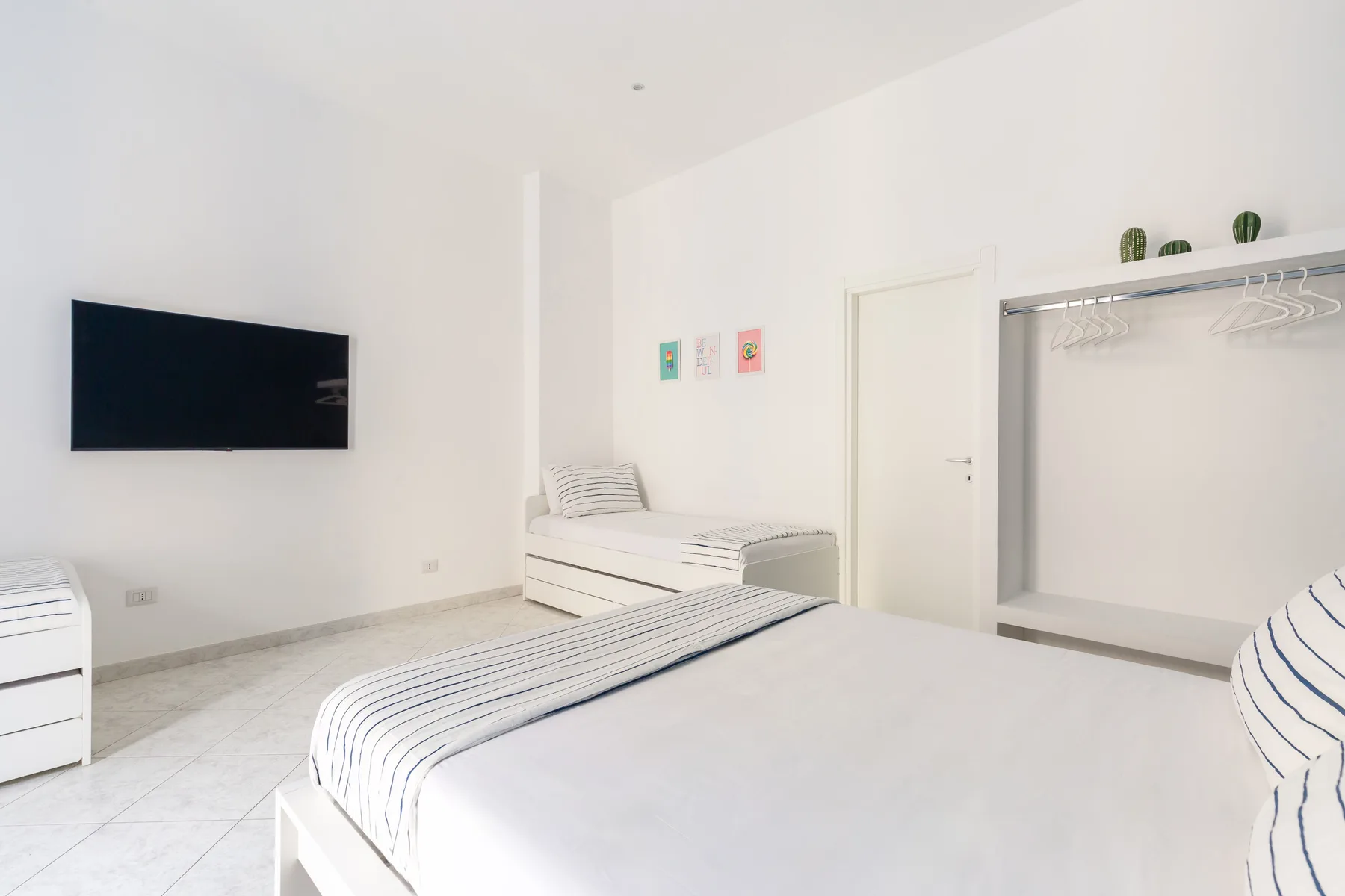 A bright bedroom with white beds, a wall-mounted television, and an open clothes rack.