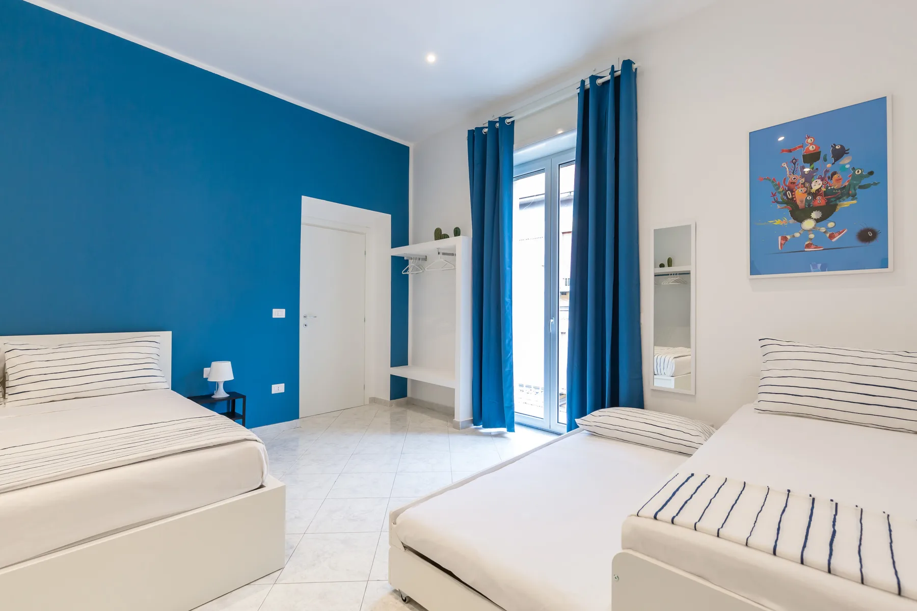 A bright bedroom with two beds and blue curtains beside a glass door.