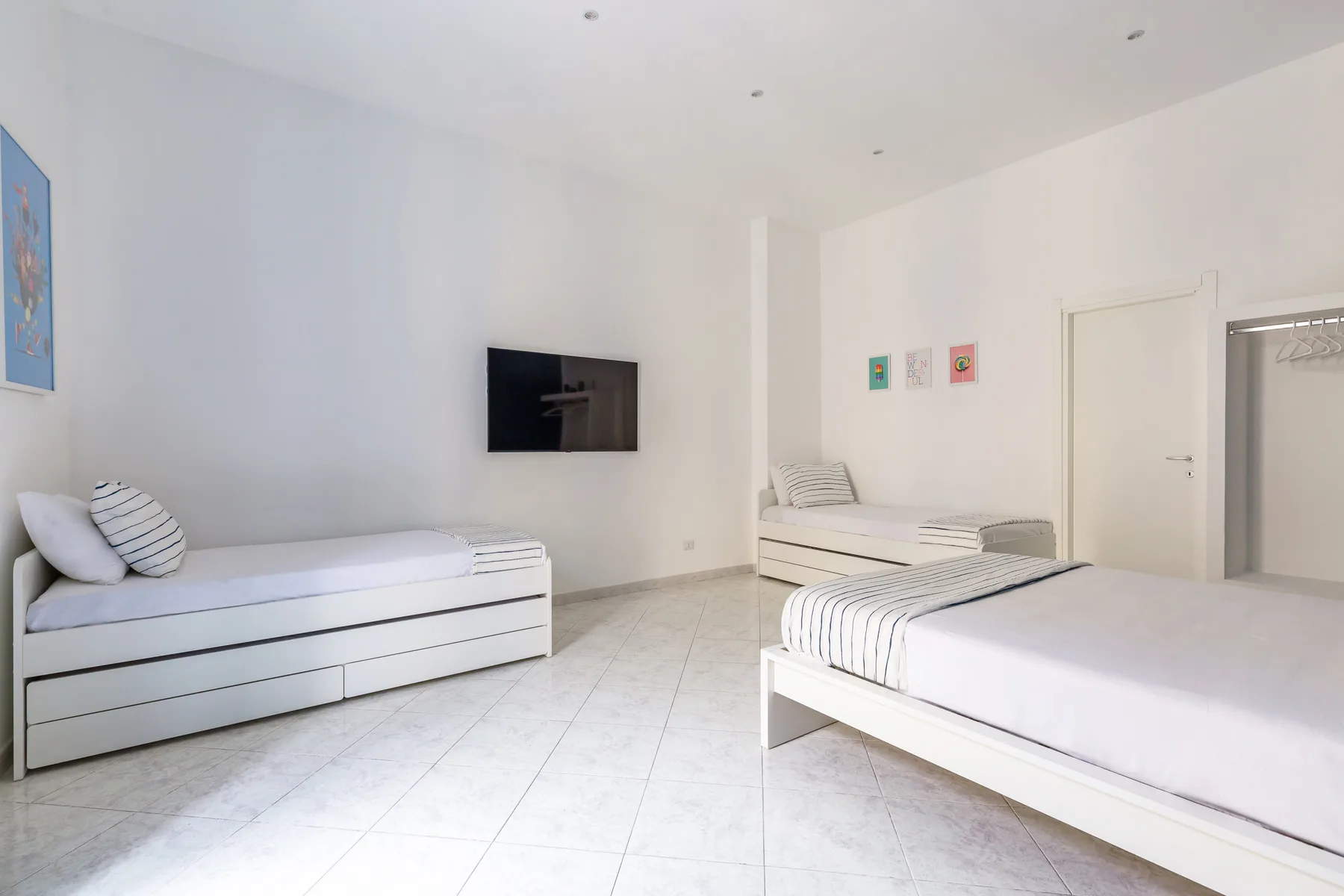 A bright bedroom with white beds, a wall-mounted television, and simple artwork.