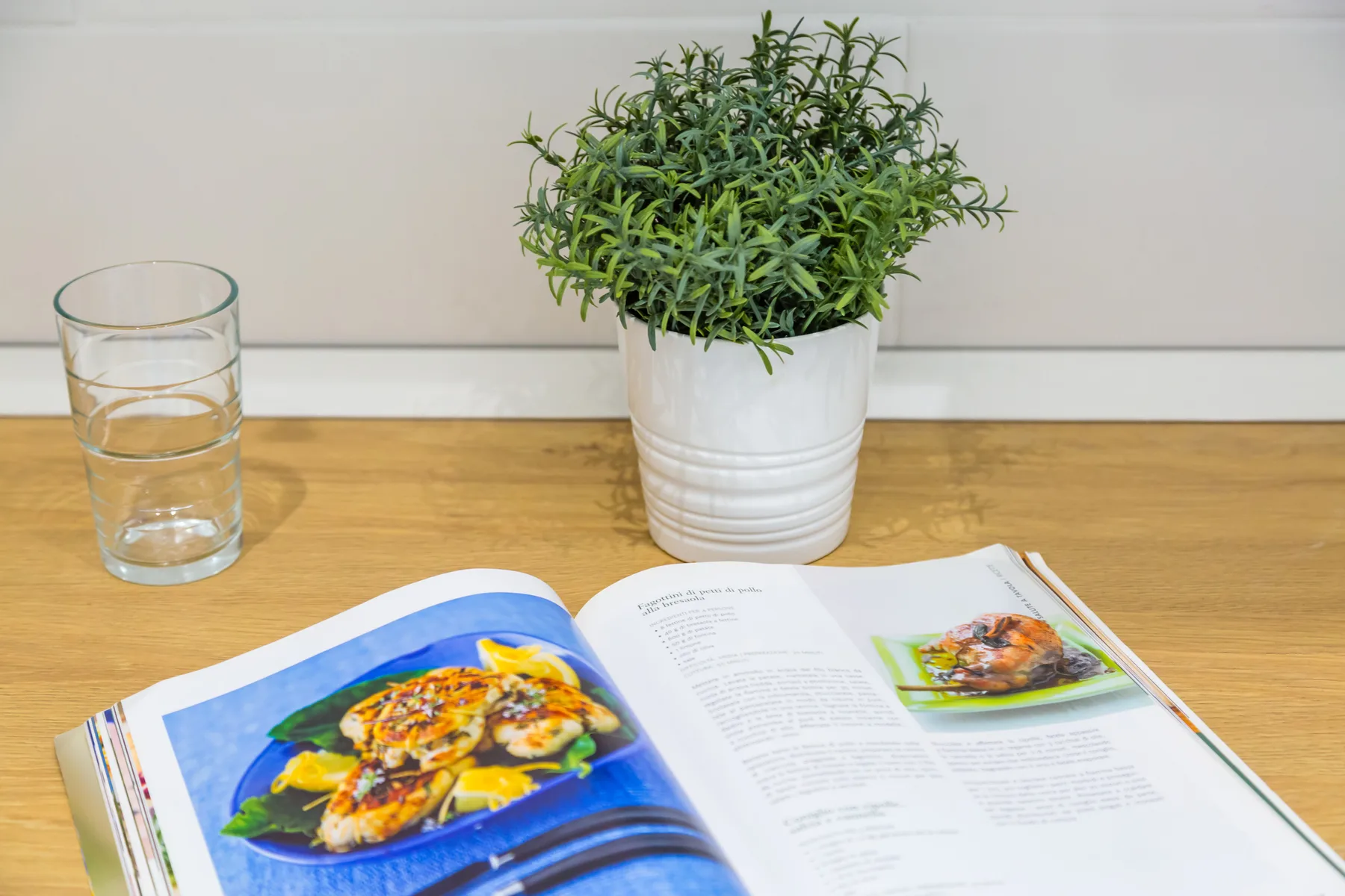 An open cookbook lies on a wooden countertop beside a glass and potted plant.
