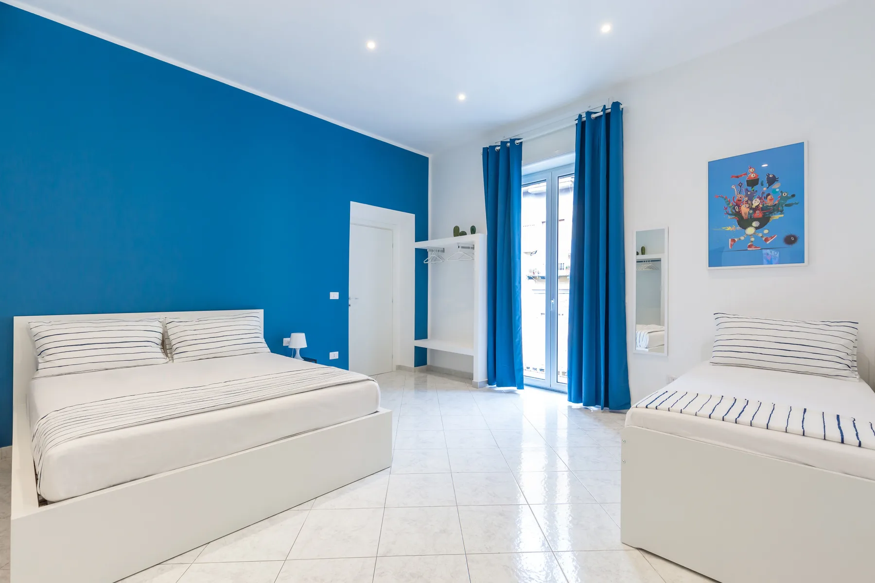 A bright bedroom with two beds, blue curtains, and a blue accent wall.