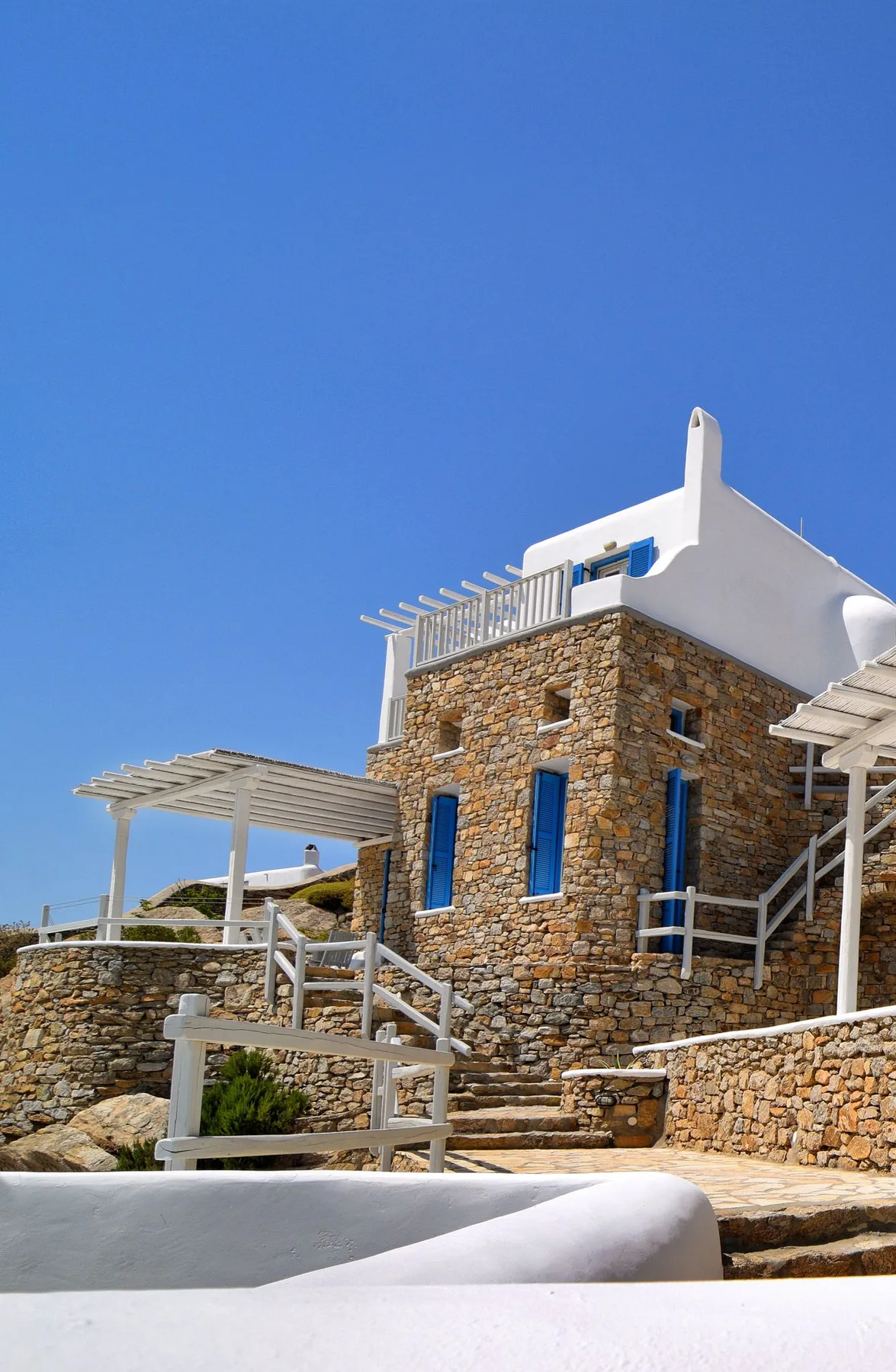 A stone building with blue windows and a white rooftop terrace beneath a clear sky.