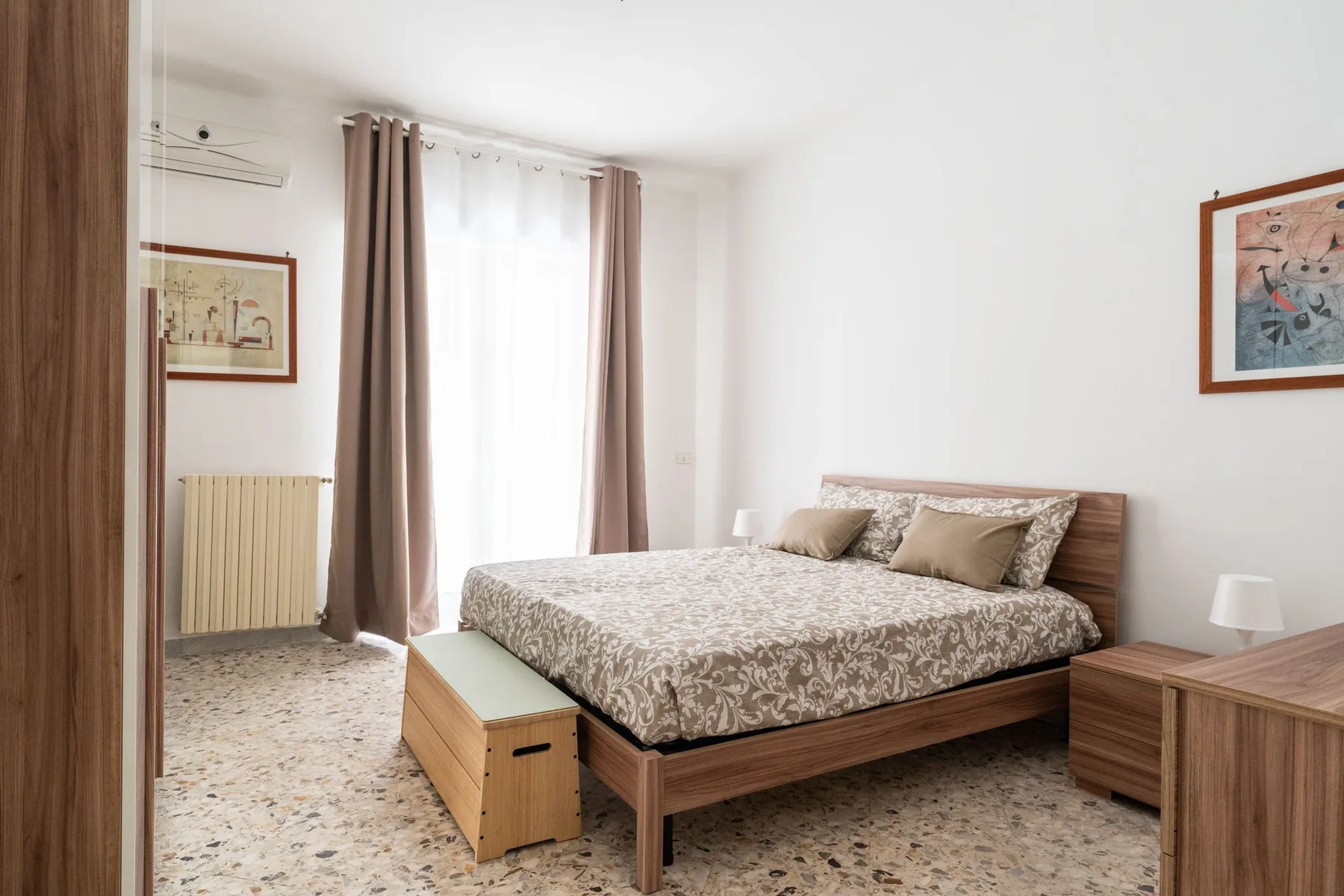 A bright bedroom with a double bed, bedside furniture, and a large curtained window.