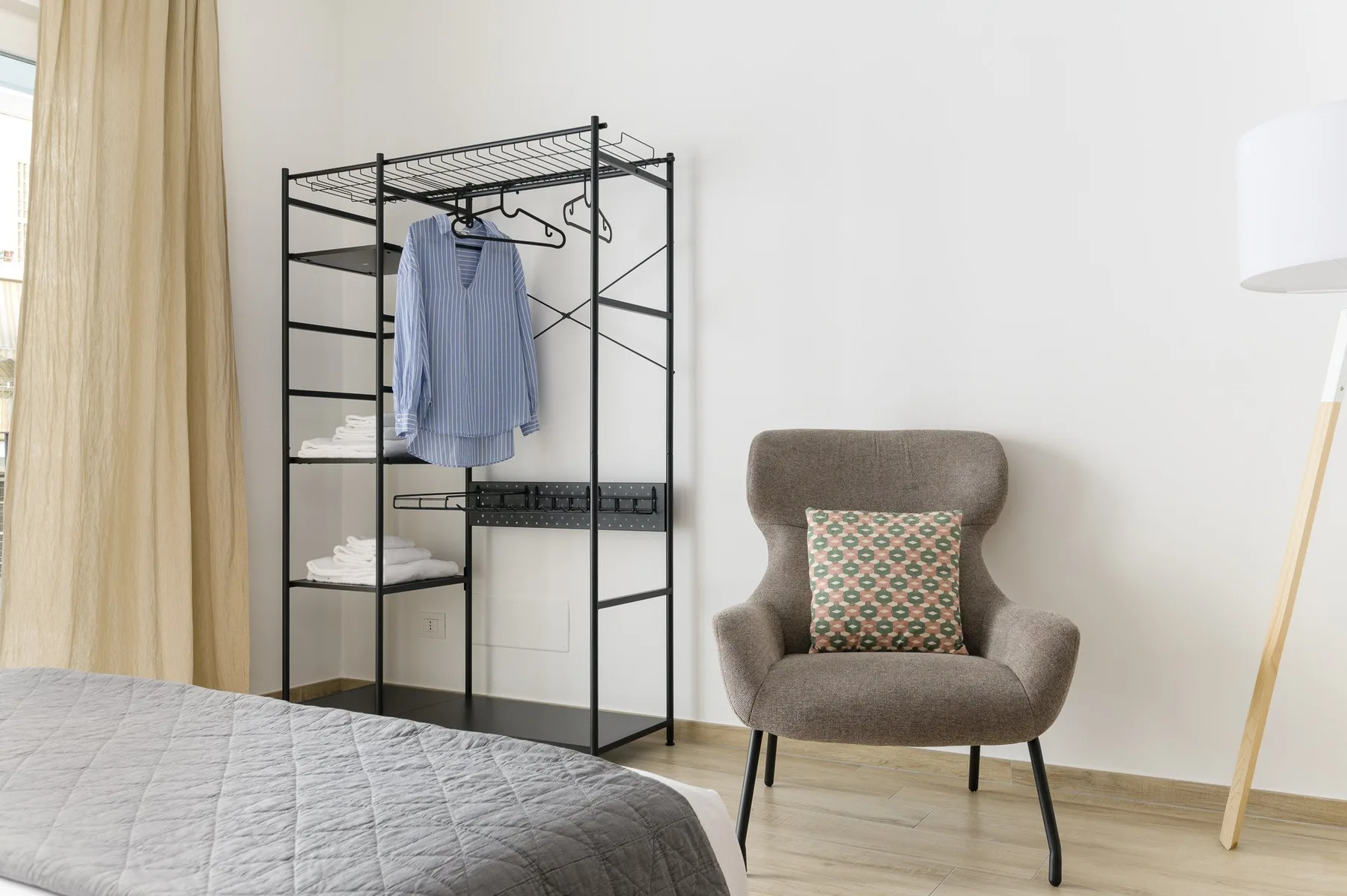 A bedroom with a clothing rack and an upholstered chair beside the bed.