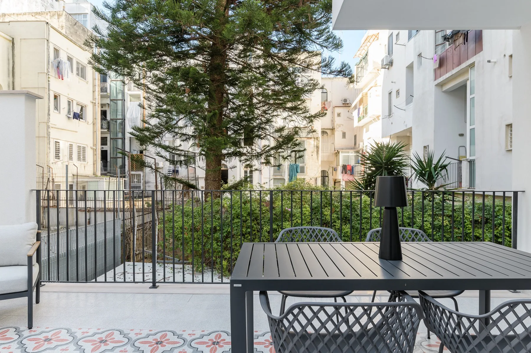 A tiled outdoor seating area overlooks a garden courtyard surrounded by apartment buildings.