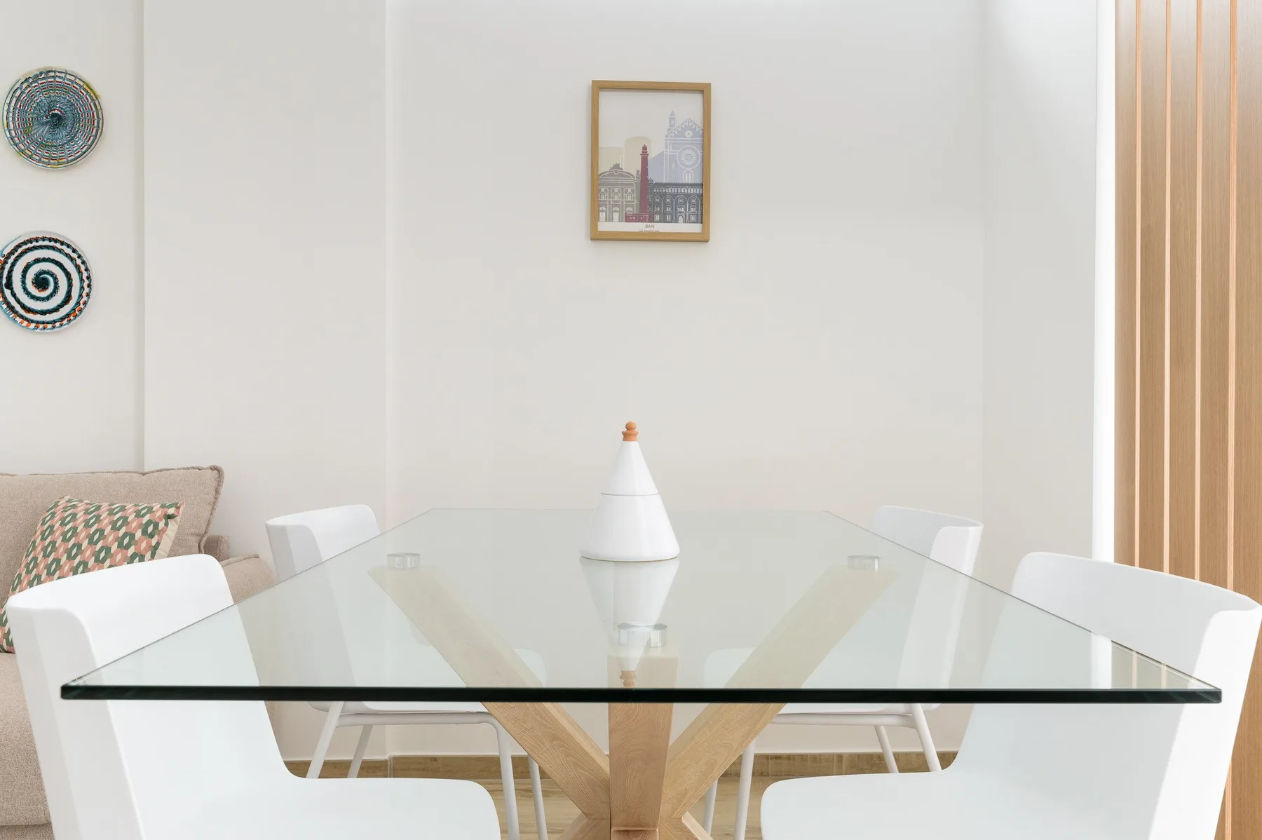 A dining area with a glass table, white seating, and framed wall art.