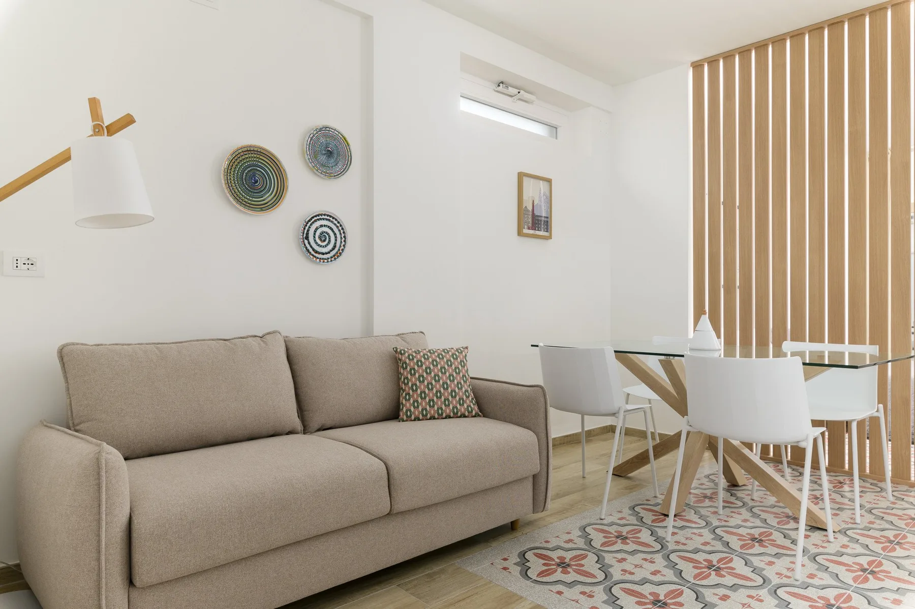 A living and dining area with a beige sofa, white table, and patterned rug.