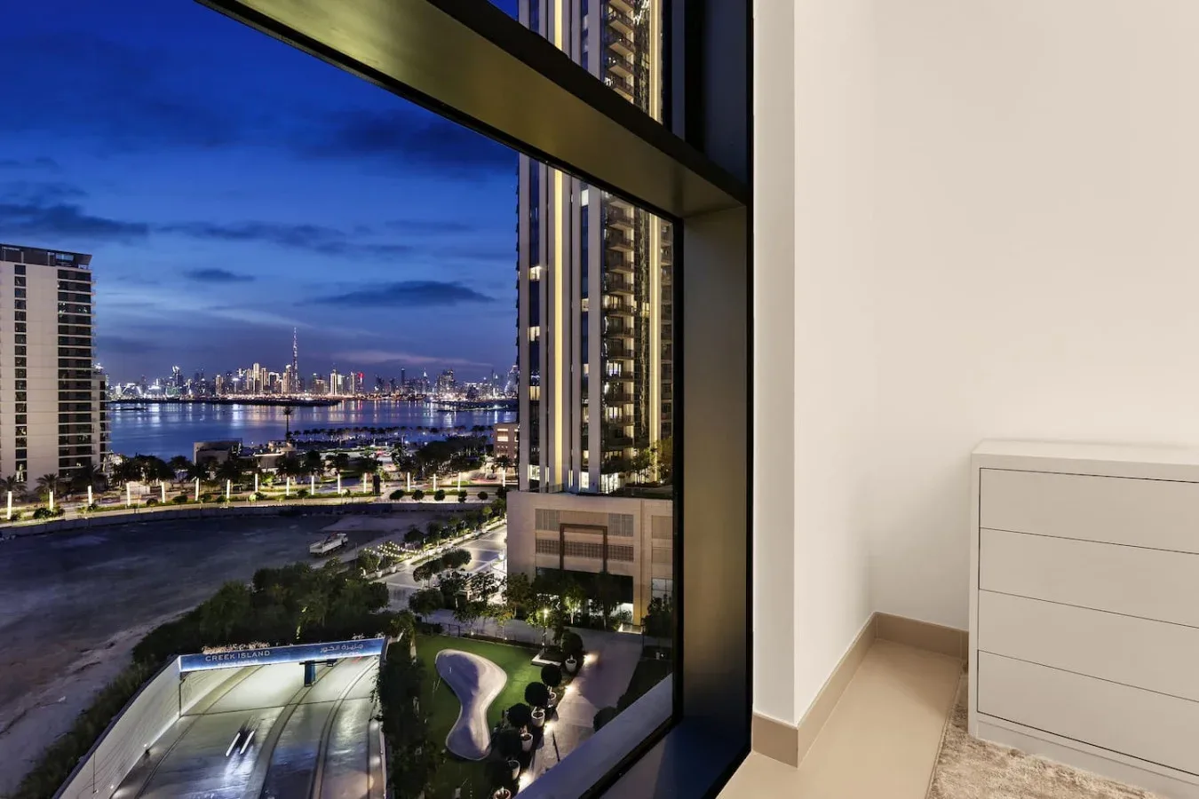 A high-rise room overlooks illuminated city buildings and a waterfront at dusk.