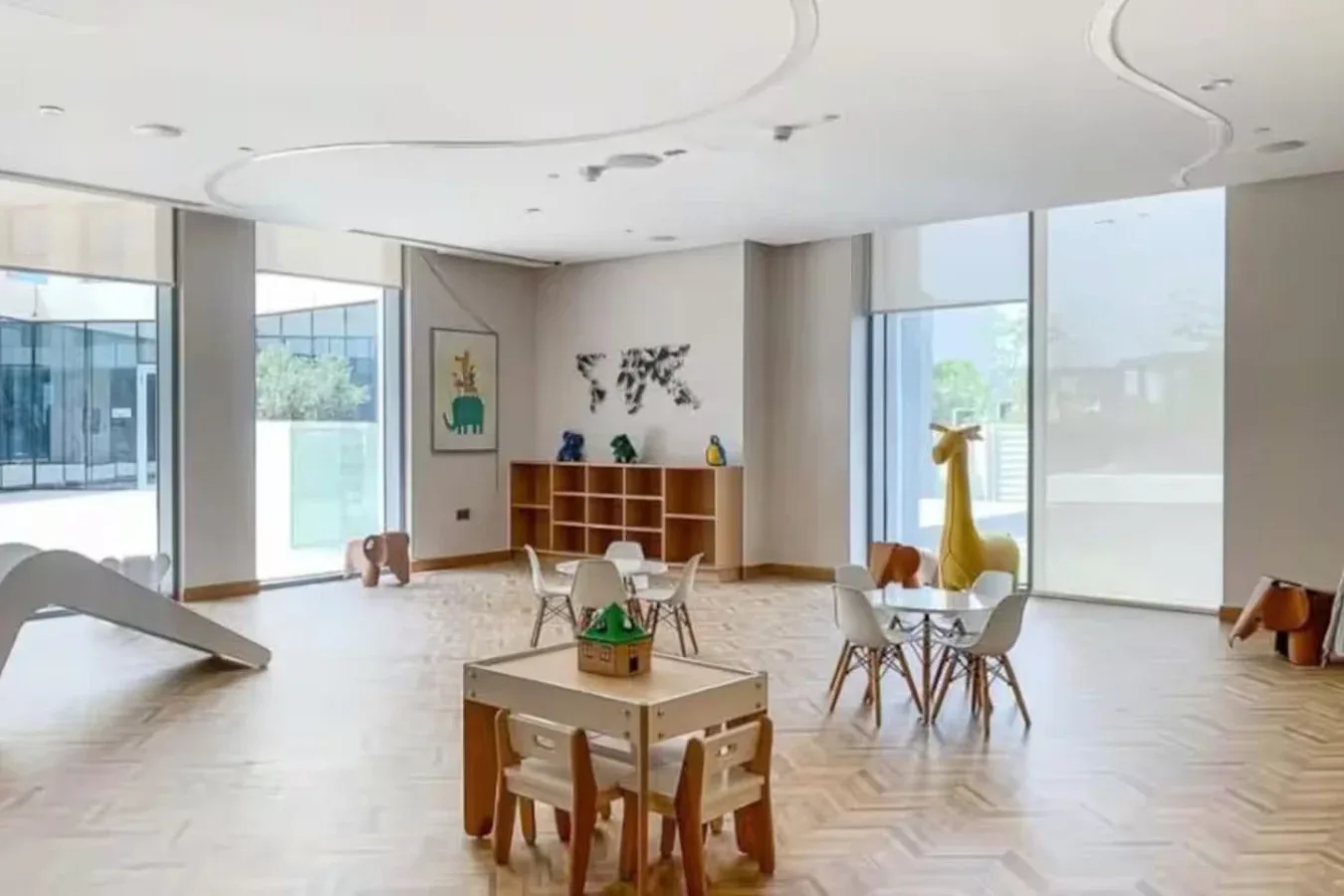 A bright playroom has small tables and chairs beside large windows.