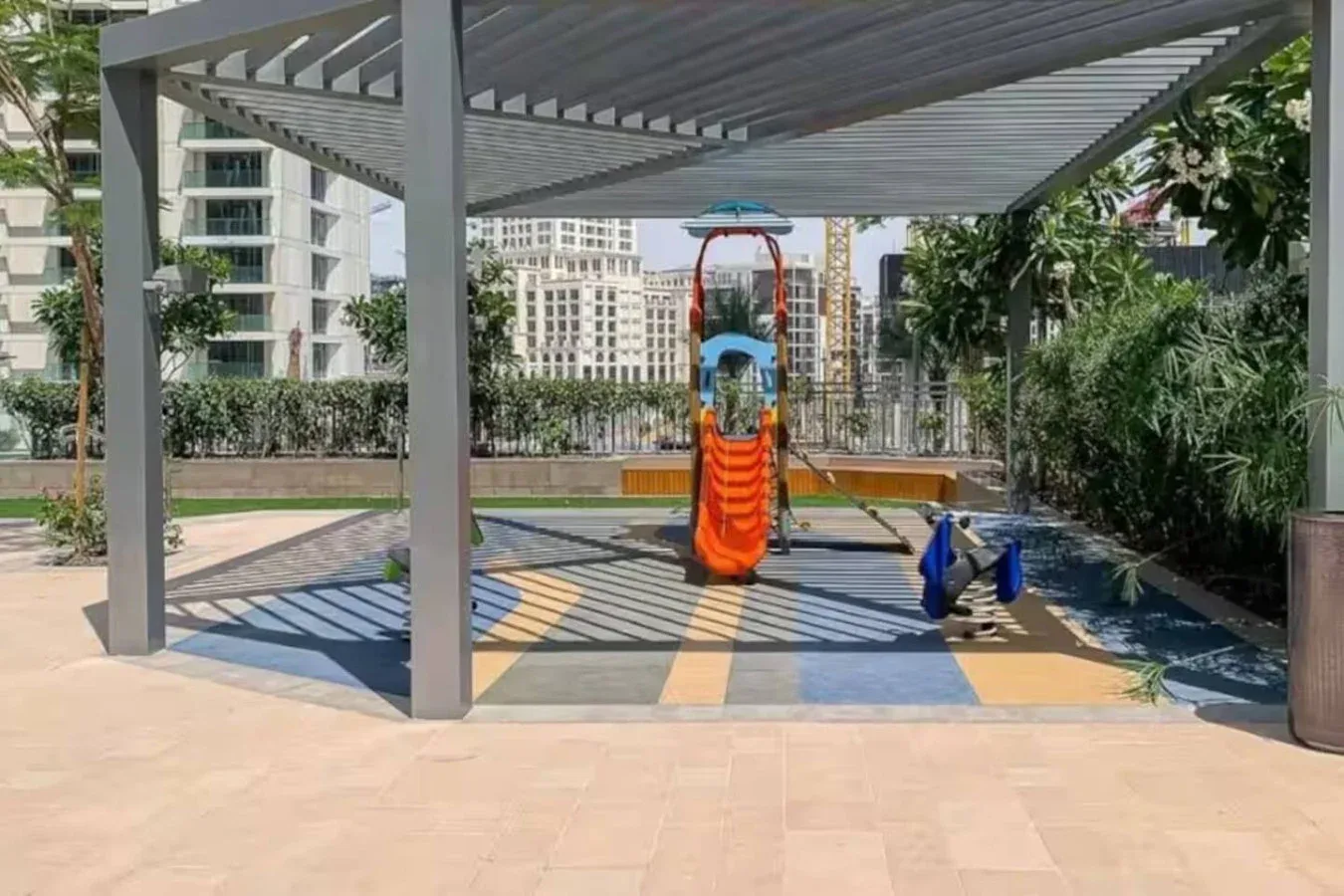 A shaded outdoor playground features colorful play equipment and rubber flooring.