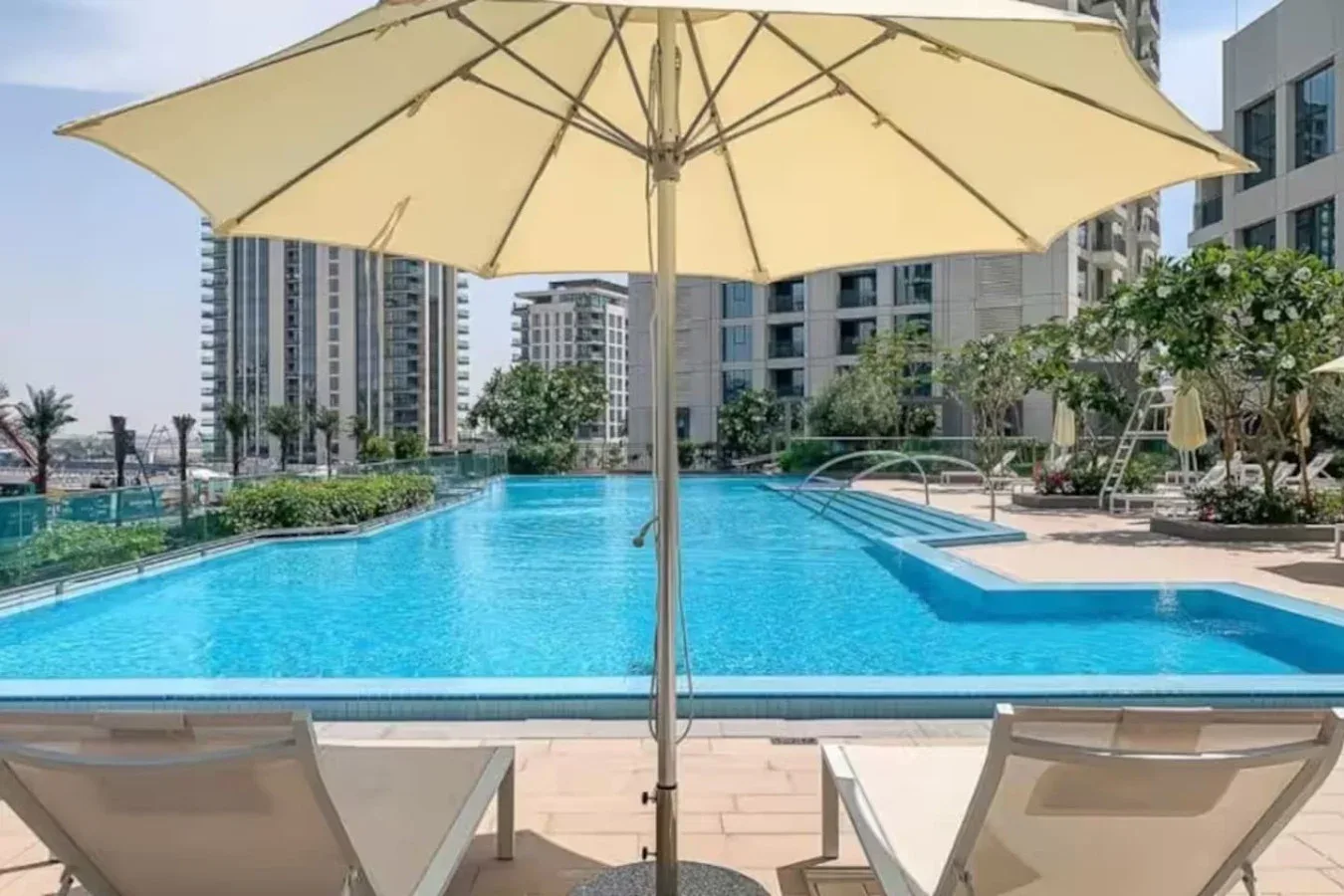An outdoor swimming pool sits among modern buildings, with loungers beneath a large umbrella.