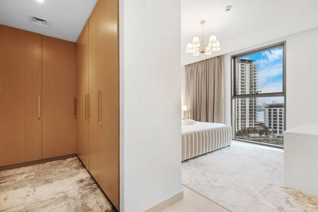 A bedroom with a bed beside a large window overlooking nearby buildings.