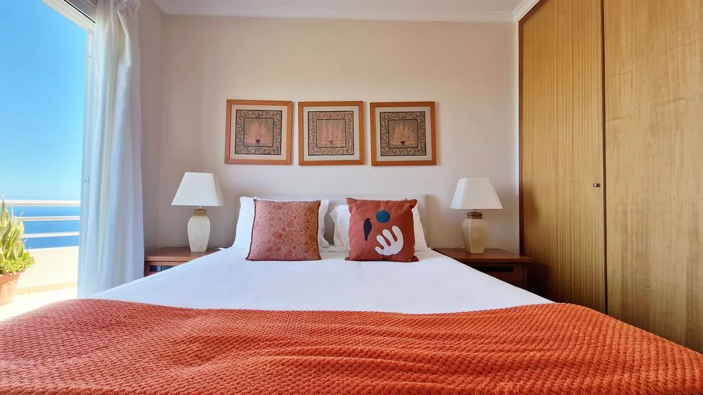Un dormitorio con una cama, cuadros enmarcados y vistas al mar a través de una puerta abierta.