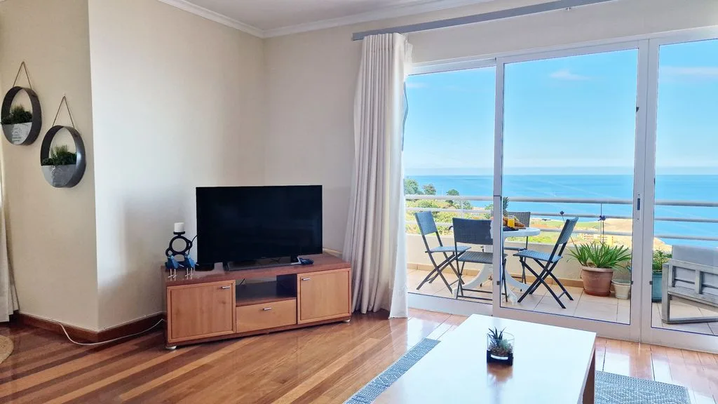 Sala de estar con televisor y zona de descanso junto a un balcón con vistas al océano.