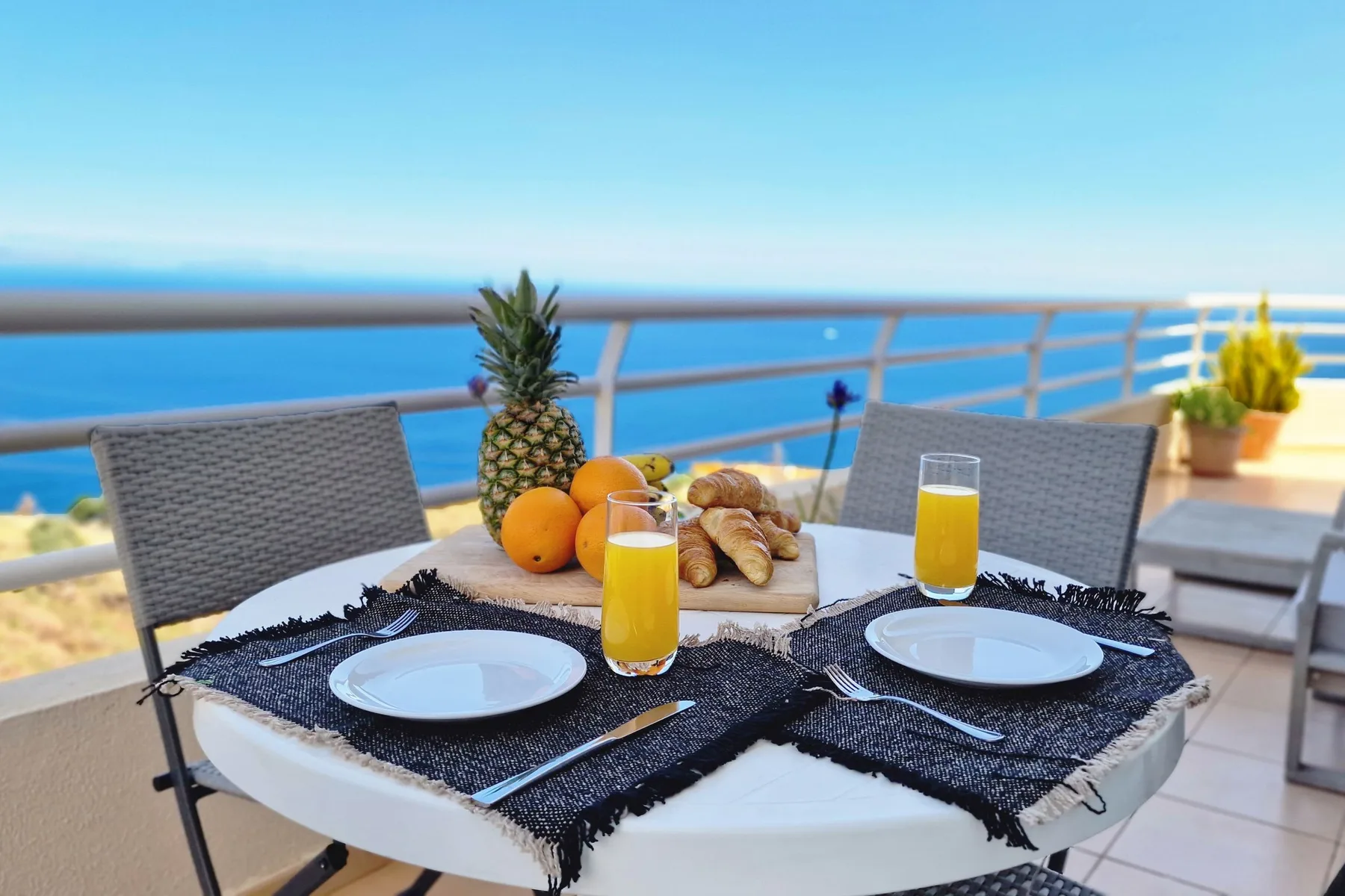 Una mesa de comedor en el patio tiene platos, bollería, fruta y zumo junto al mar.