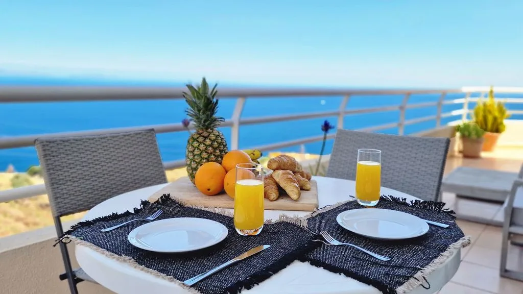 Una mesa de comedor en un balcón está puesta con platos, fruta, pan y bebidas frente al mar.