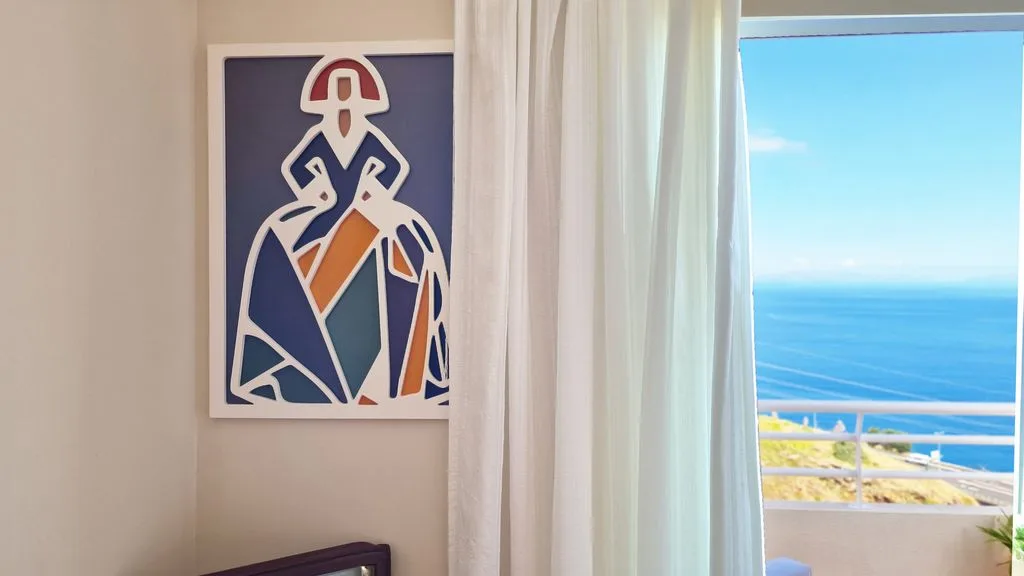 Una habitación luminosa con una obra abstracta enmarcada junto a una ventana con vistas al mar.