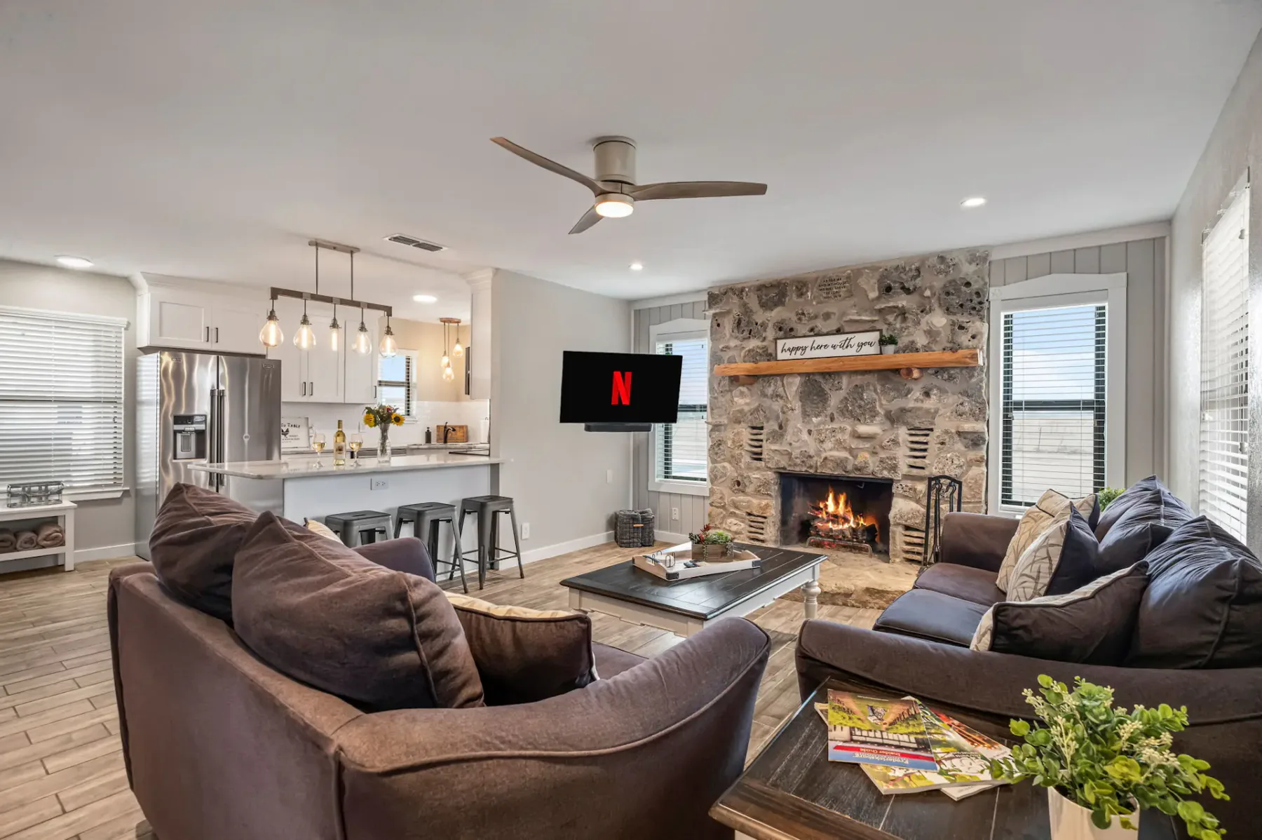 Sala de estar moderna con chimenea de piedra, televisor de pantalla plana, sofás acogedores y una cocina al fondo. Los elementos decorativos incluyen plantas y revistas.