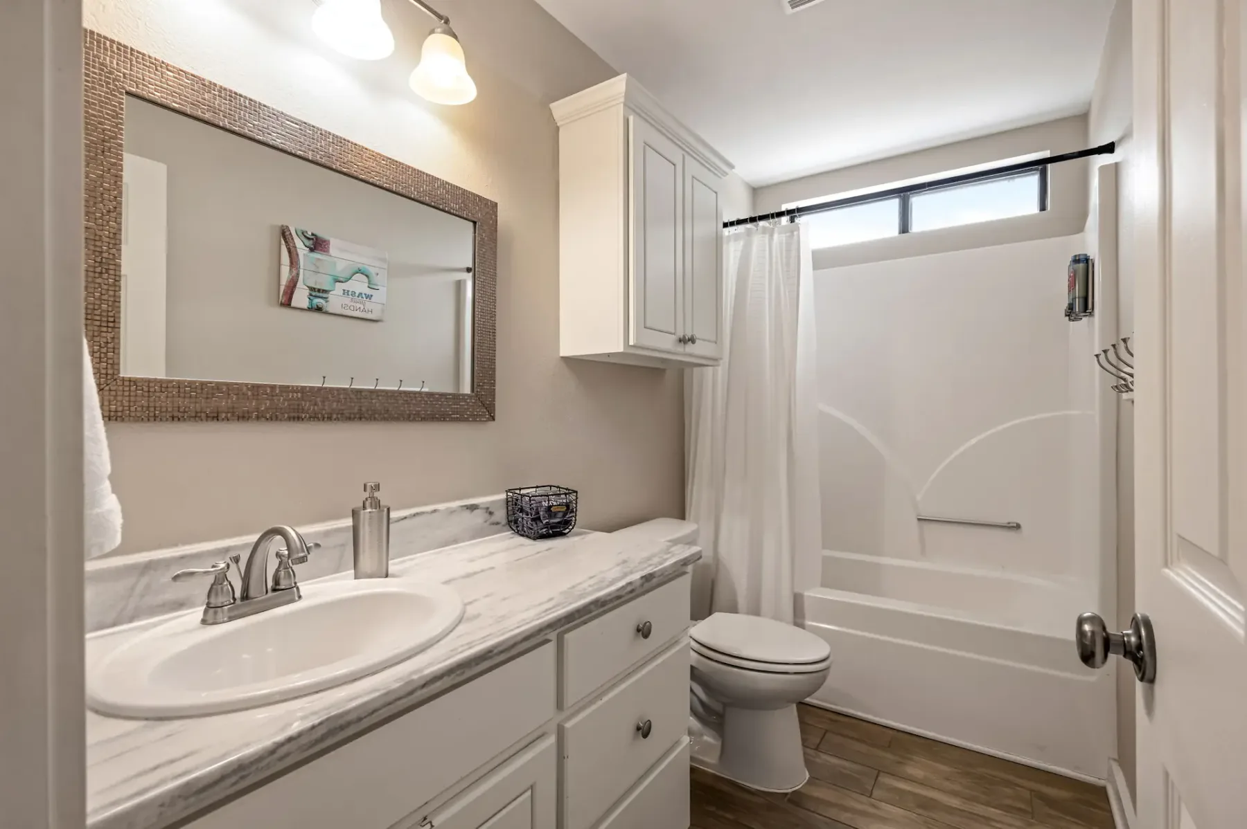Baño con un mueble blanco y encimera de mármol, espejo con marco, inodoro, bañera con cortina y suelo de madera, iluminado por luces de techo.