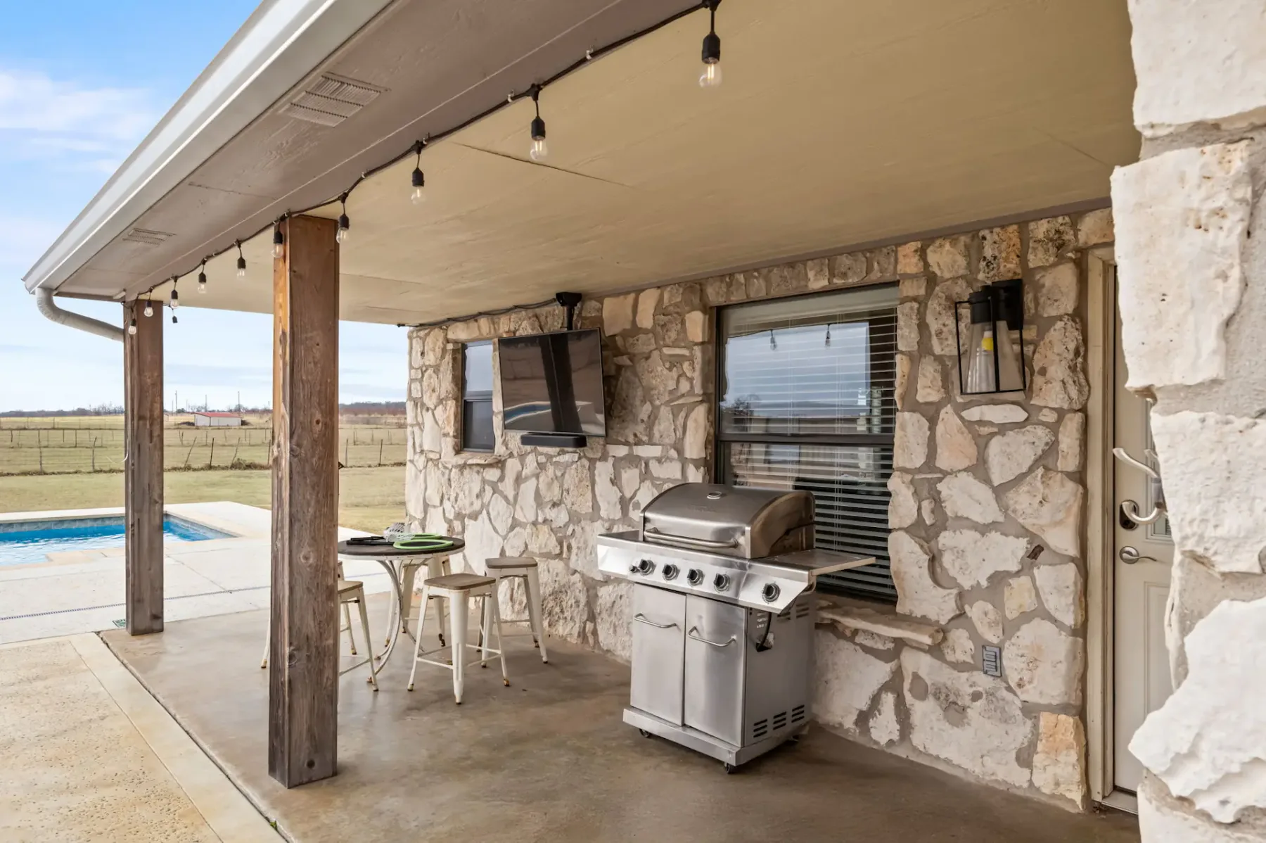 Patio cubierto con una pared de piedra, parrilla exterior, taburetes de bar y guirnaldas de luces, con vistas a una piscina y un paisaje abierto.