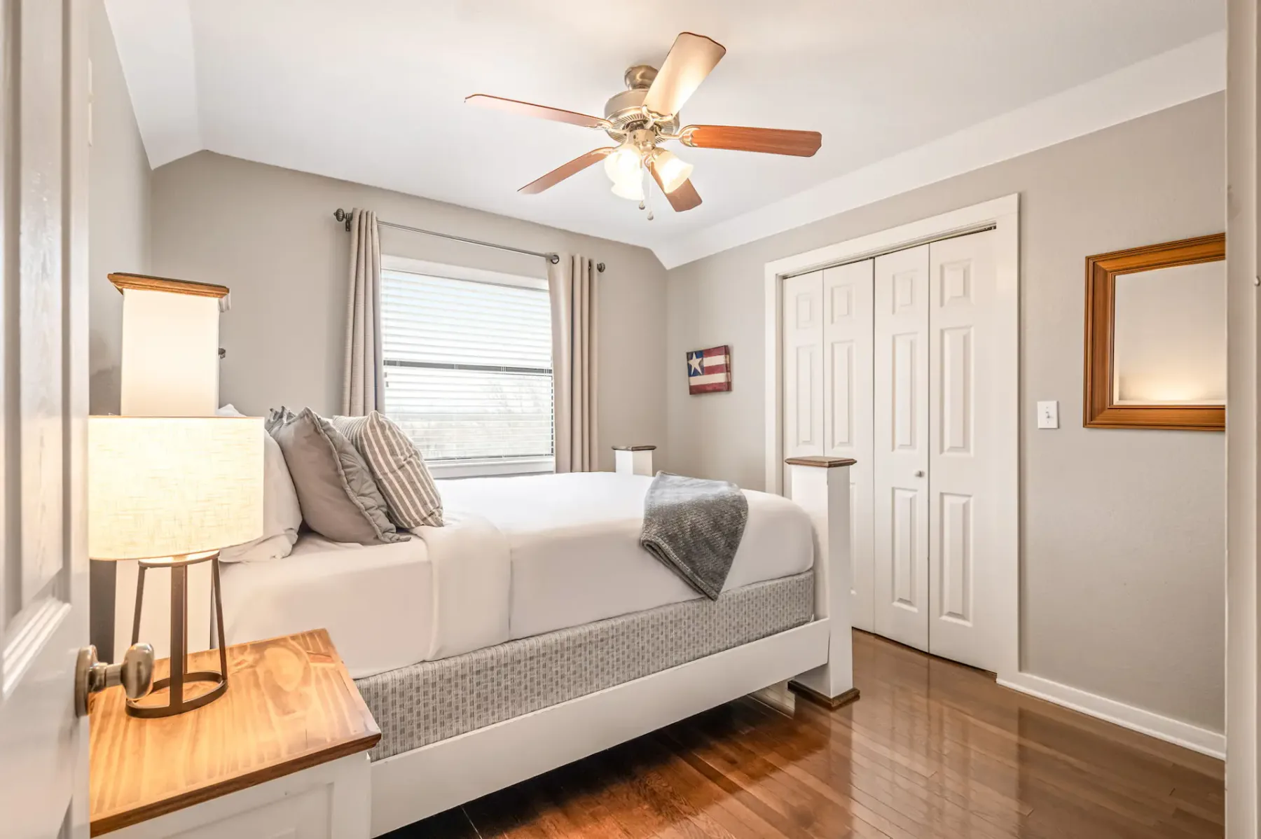 Dormitorio luminoso y moderno con una cama grande, suelo de madera, ventilador de techo y una ventana con cortinas. Se ven una lámpara de noche y un armario.