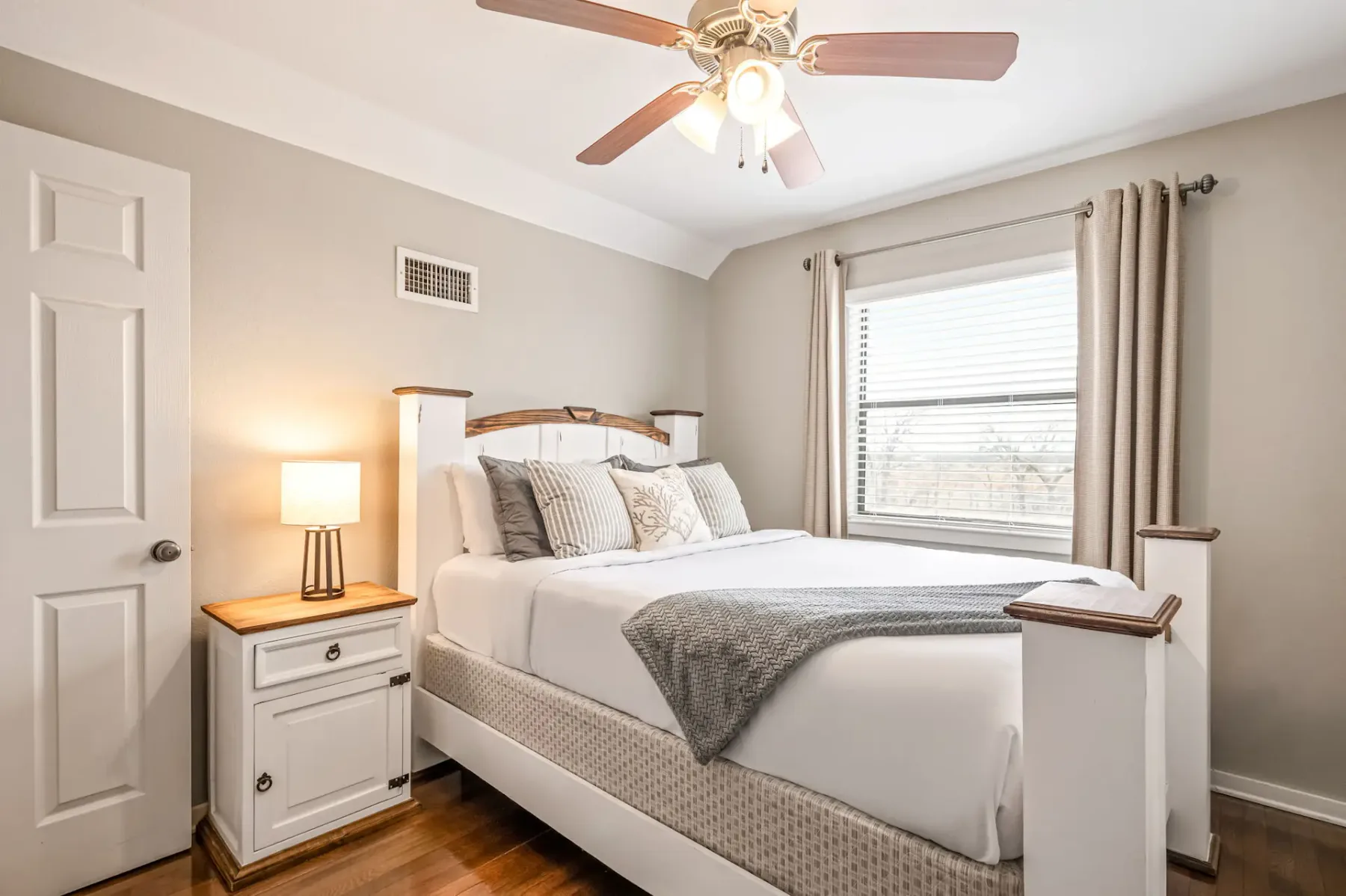Dormitorio acogedor con una cama bien tendida, cabecero de madera, mesa de noche con lámpara y ventilador de techo. La luz del sol entra por una ventana.