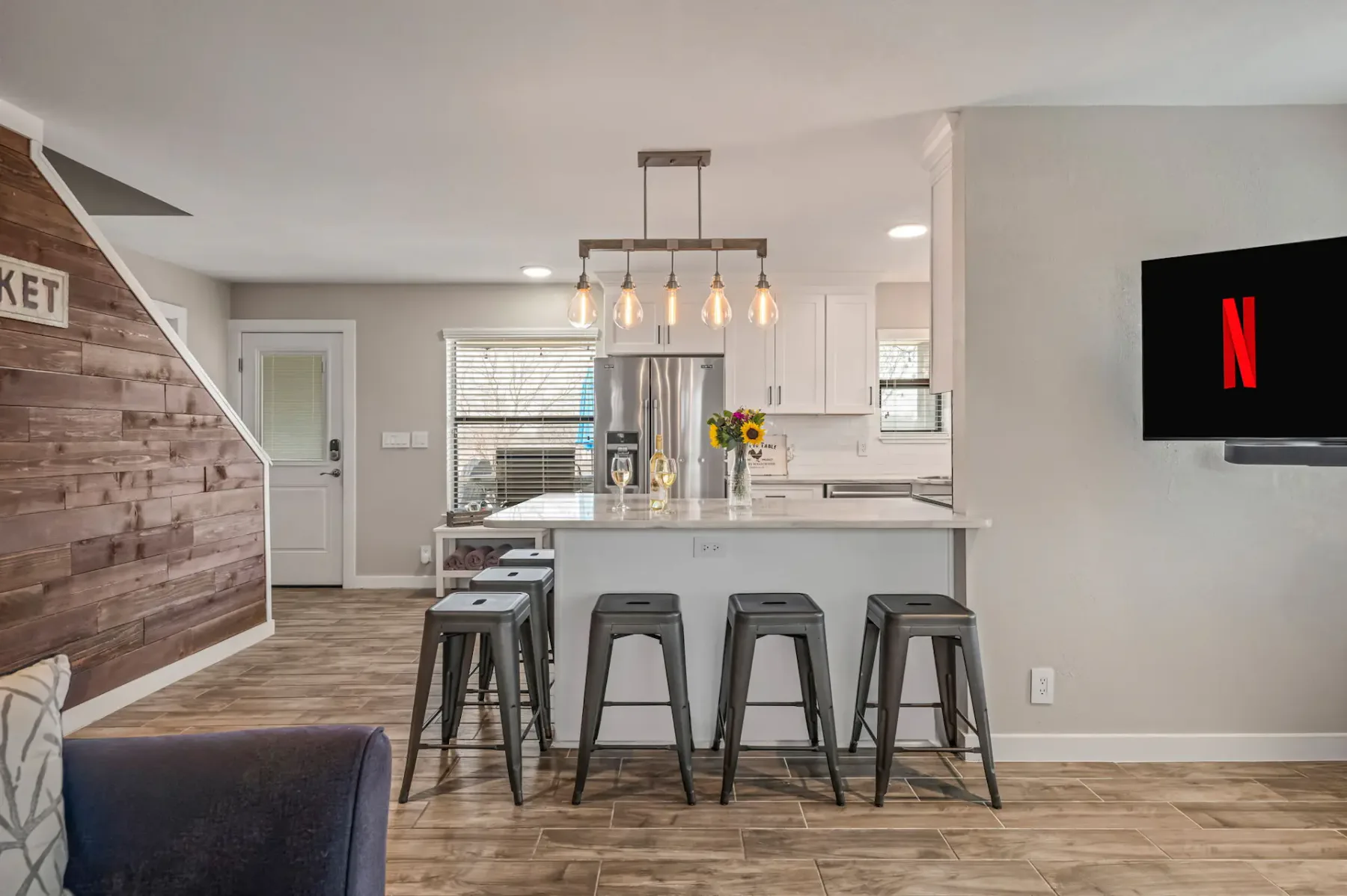 Cocina moderna con una isla central, taburetes de bar, lámparas colgantes y detalles de madera. En la pared hay un televisor con el logotipo de Netflix.