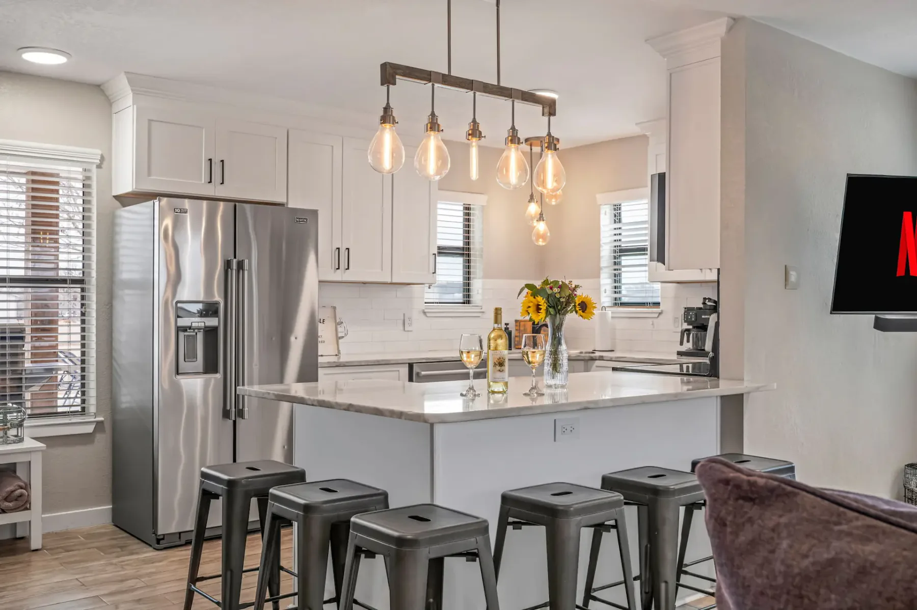 Cocina moderna con armarios blancos, una isla de mármol, lámparas colgantes y electrodomésticos de acero inoxidable. Hay taburetes de bar a lo largo de la isla.