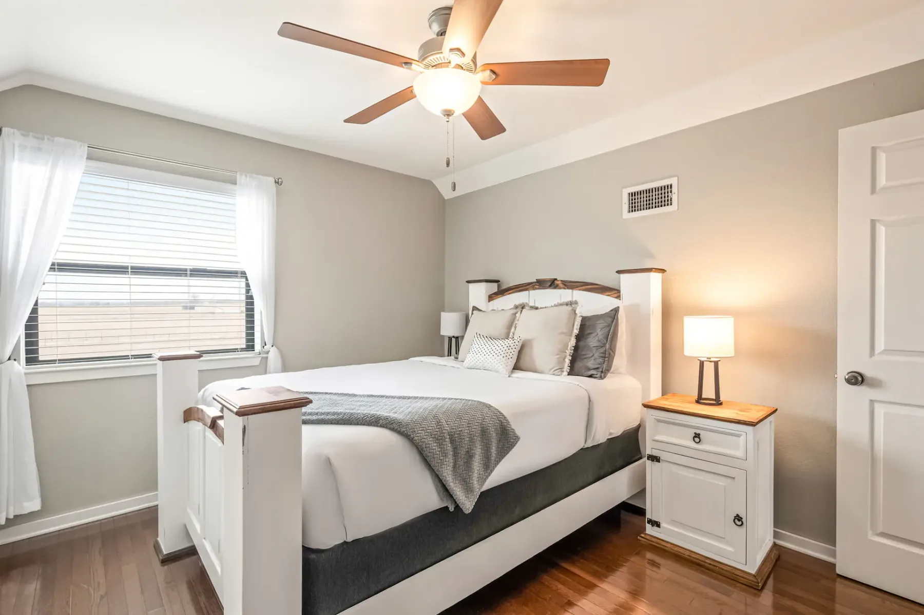 Dormitorio luminoso con una cama grande, ropa de cama blanca, una manta decorativa gris, mesas de noche con lámparas, suelo de madera, ventilador de techo y una ventana con cortinas translúcidas.