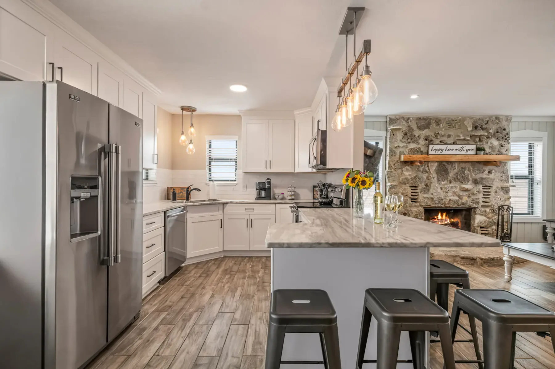 Cocina y sala de estar modernas de concepto abierto con techos altos, lámparas colgantes, un sofá beige, una mesa de comedor de madera y electrodomésticos de acero inoxidable.