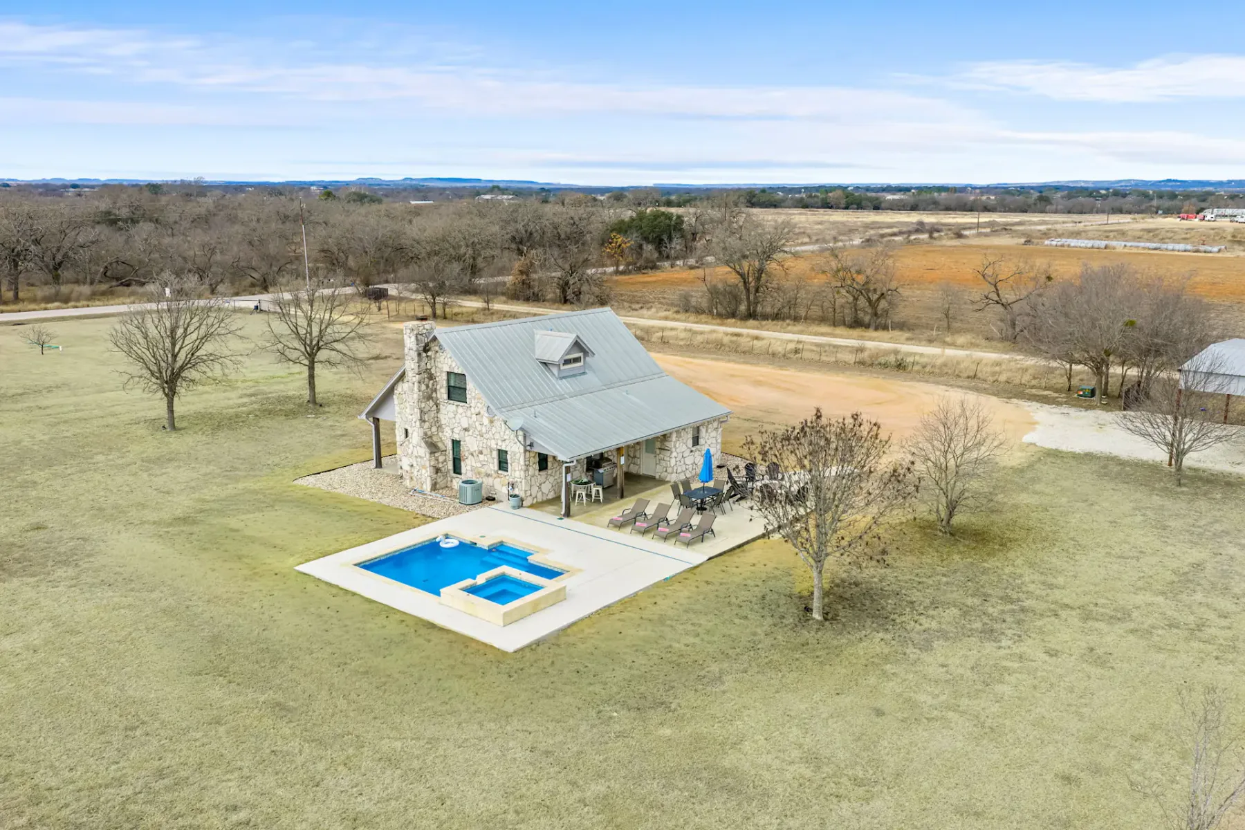 Casa de piedra con techo de metal, rodeada de campos abiertos, con piscina y jacuzzi en el patio trasero, asientos al aire libre y árboles dispersos.