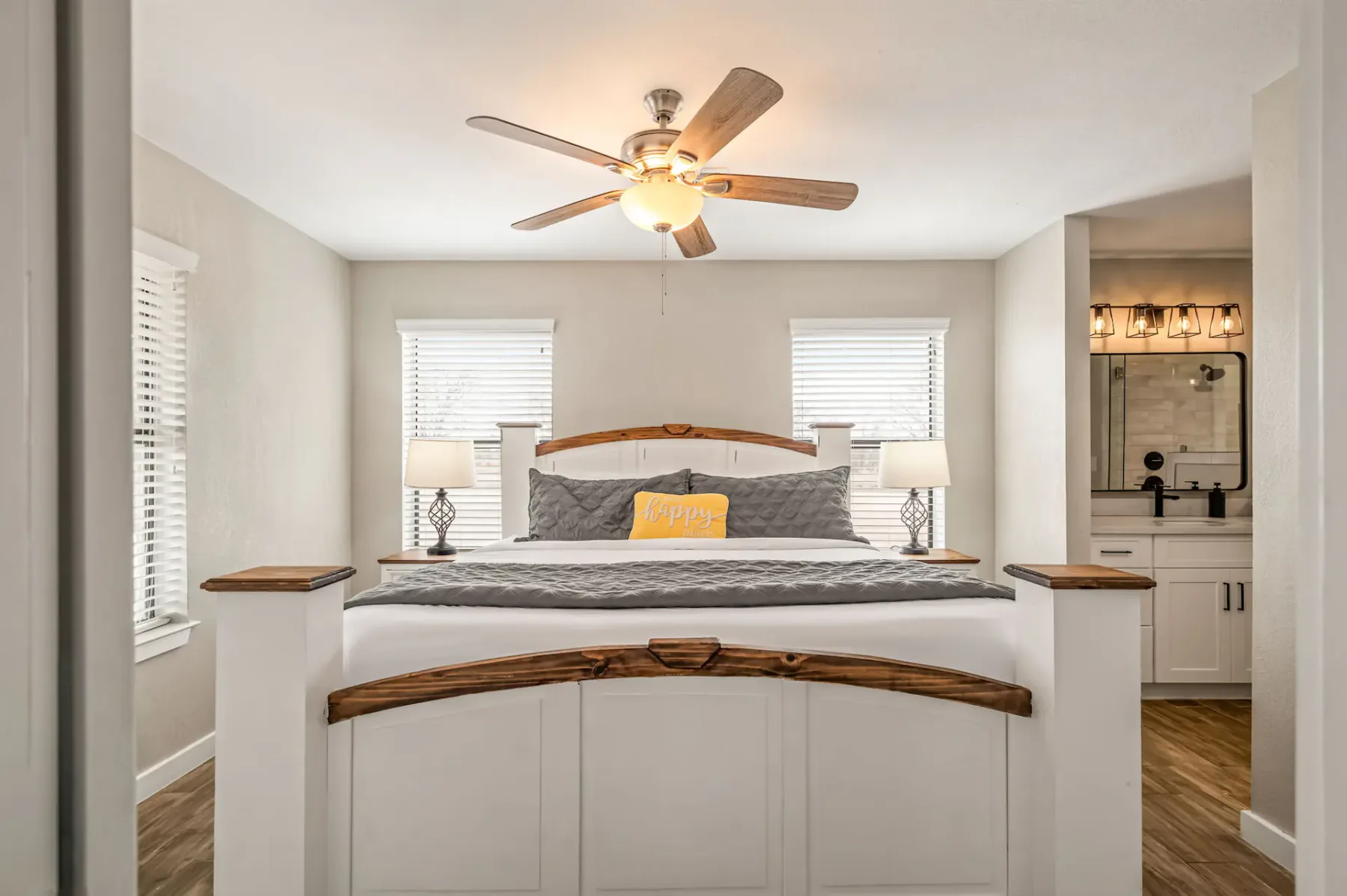 Un dormitorio acogedor con una cama king size, dos lámparas de noche y un ventilador de techo. La habitación tiene paredes claras, suelo de madera y una pequeña zona de baño.