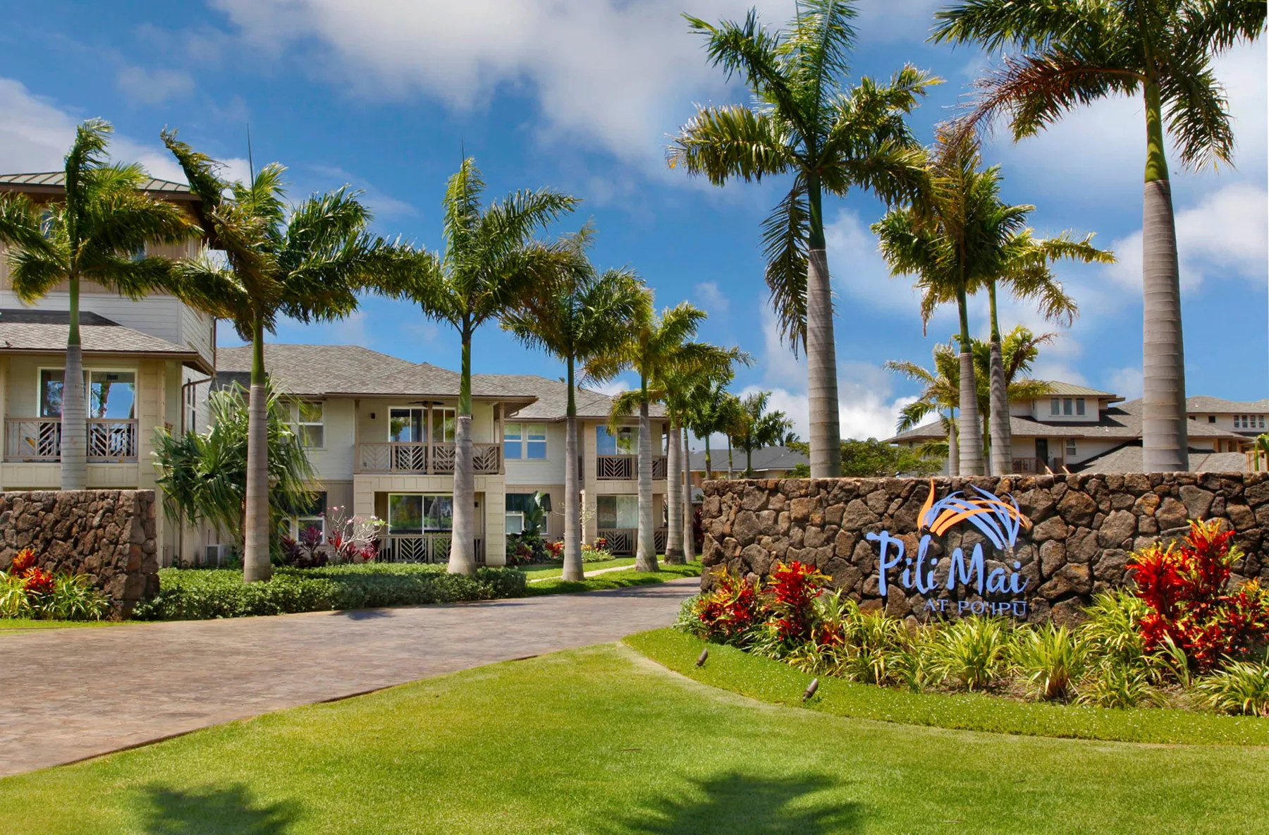 A tropical resort entrance with palm trees, landscaped gardens, and a stone wall.