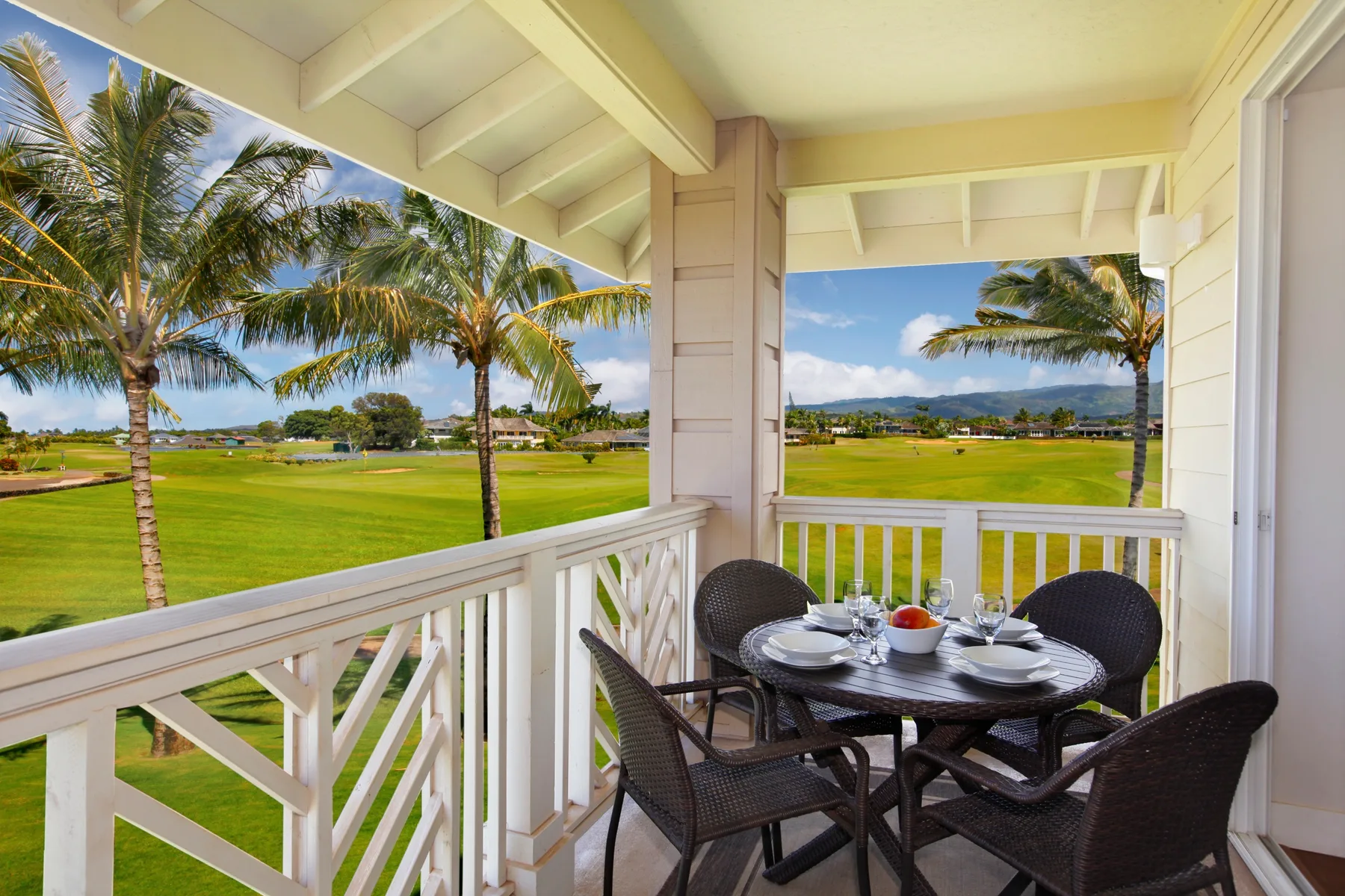 A covered balcony with outdoor seating overlooks a green lawn and palm trees.
