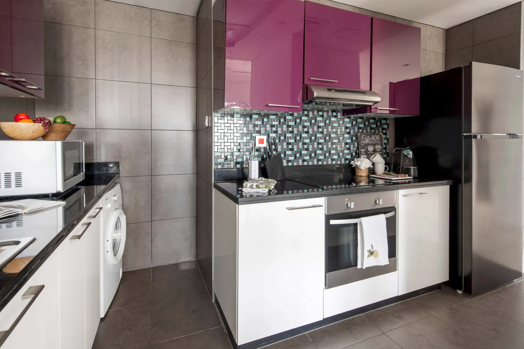 Una cocina compacta con gabinetes blancos, alacenas superiores magenta y un salpicadero de azulejos.