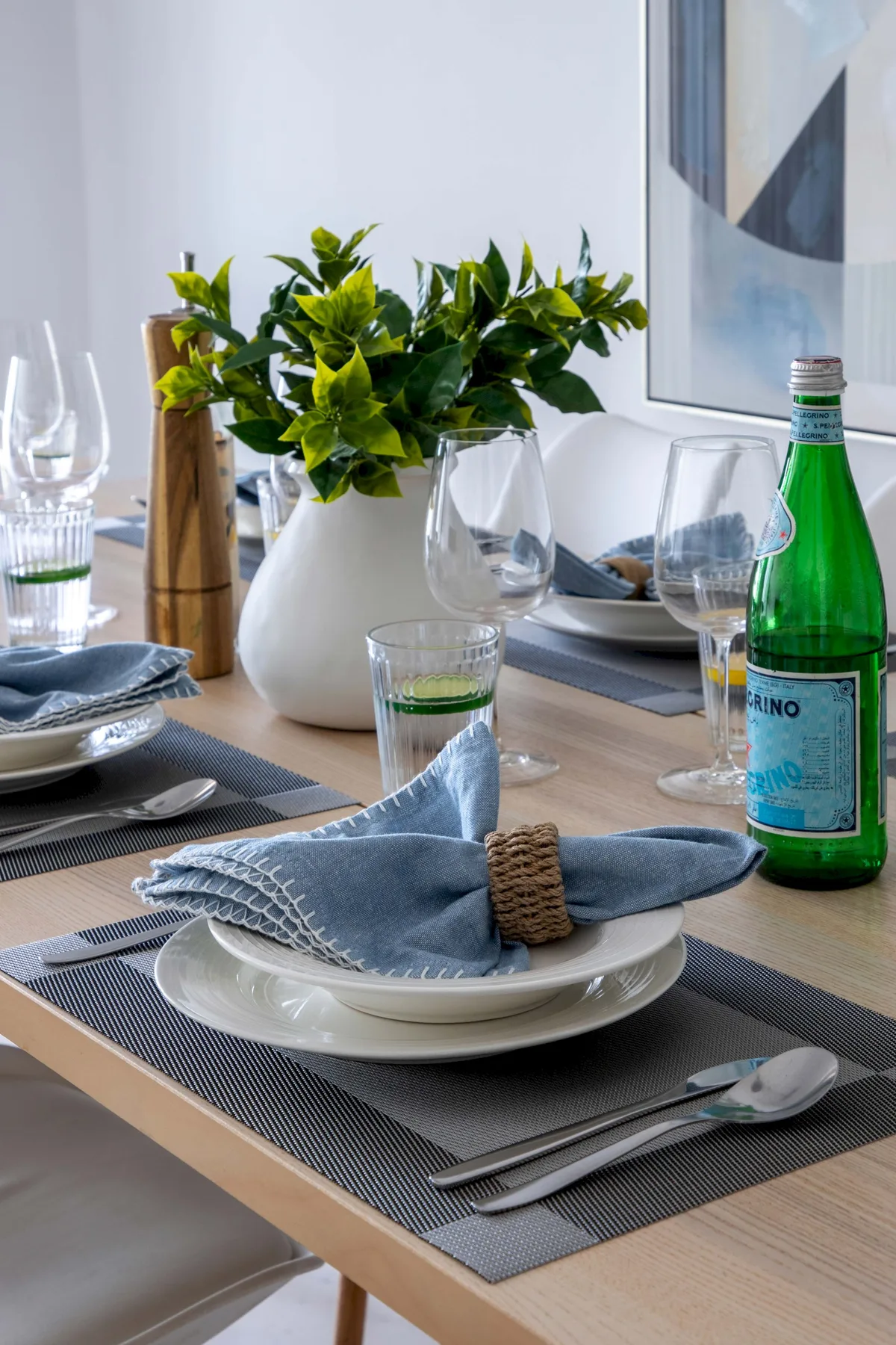 Una mesa de comedor está preparada con platos, servilletas dobladas, cristalería y un centro de mesa con hojas.