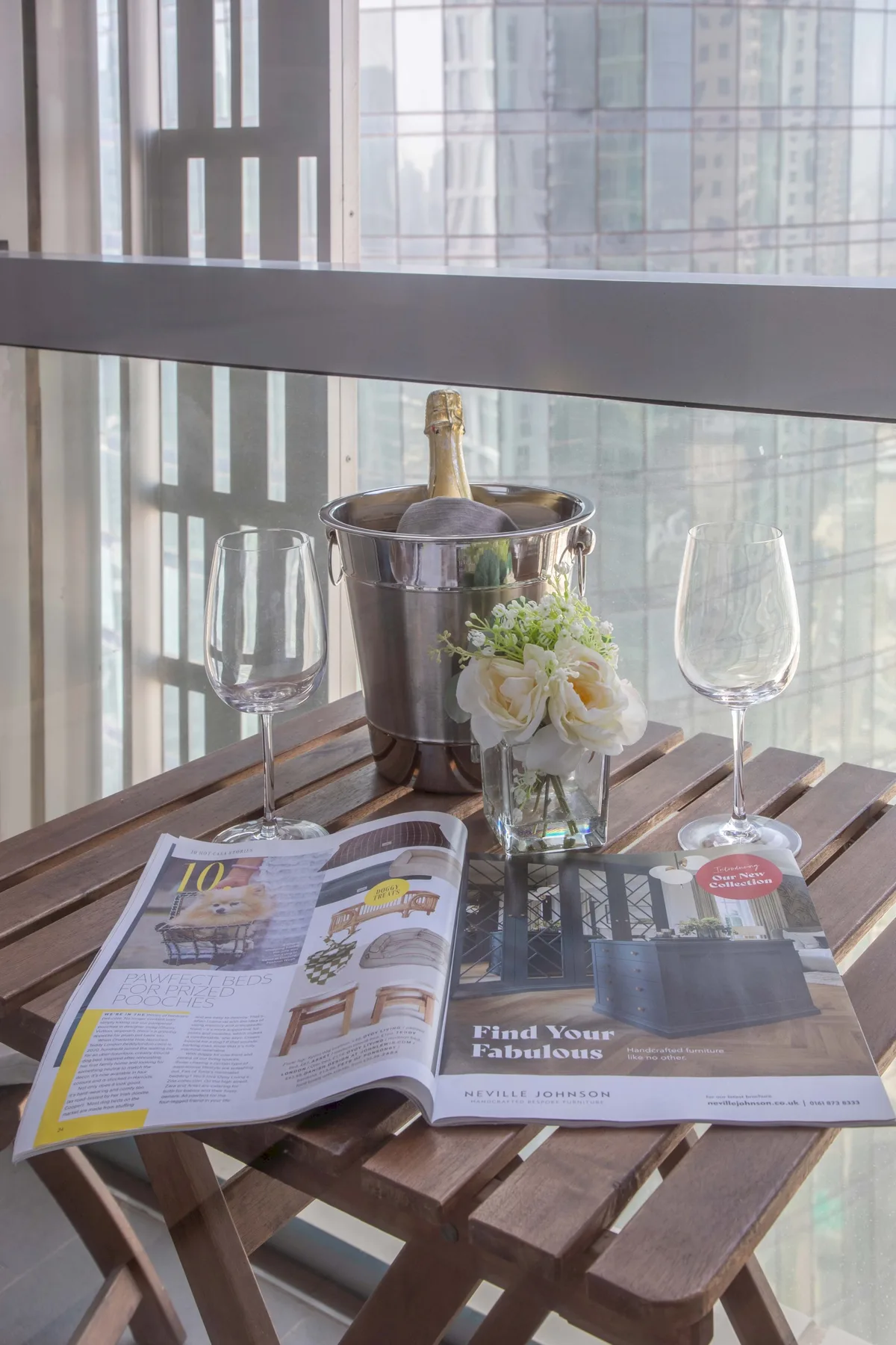 Una mesa pequeña junto a una ventana tiene una revista abierta, flores y una cubitera.