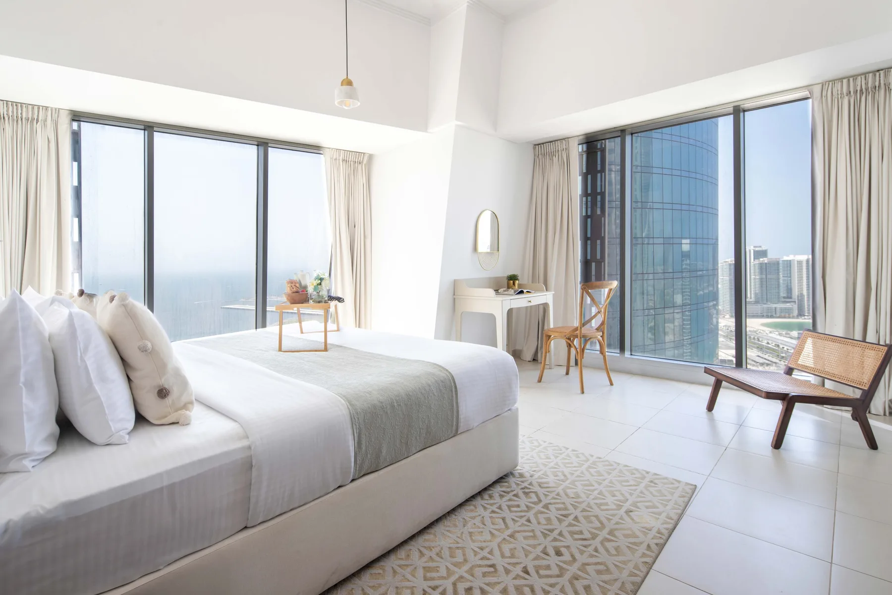 Un dormitorio luminoso con una cama junto a grandes ventanales con vistas a edificios.