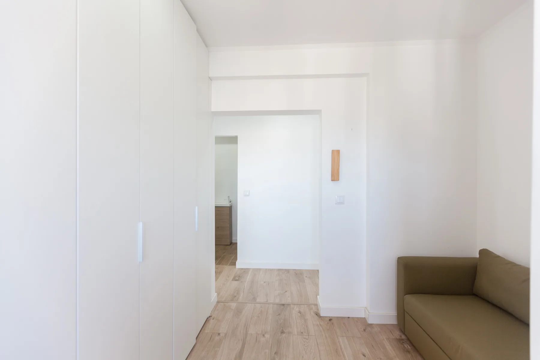 A bright hallway opens toward a room with light wood flooring and seating.