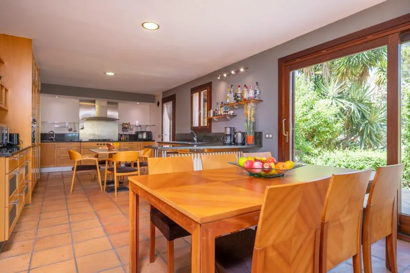 A dining area with a wooden table beside a bright kitchen and large window.