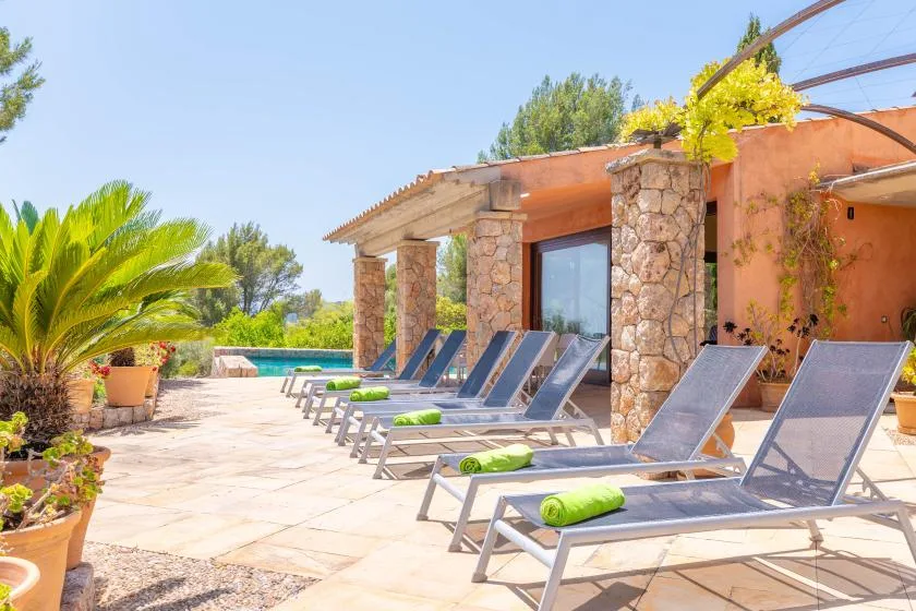 A sunny poolside patio has rows of loungers beside a stone-columned building.