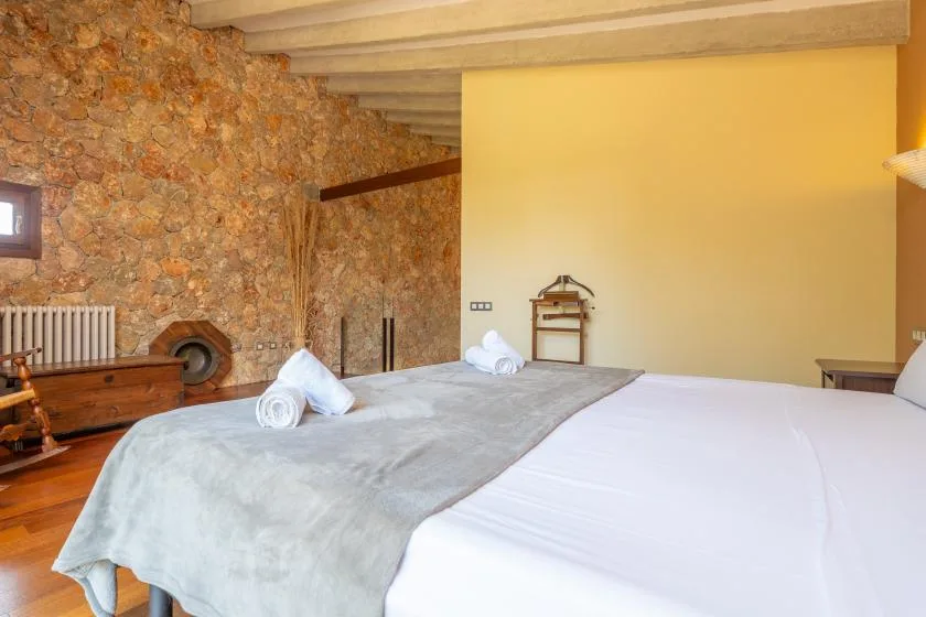 A bedroom with a large bed, pale bedding, and a stone wall.